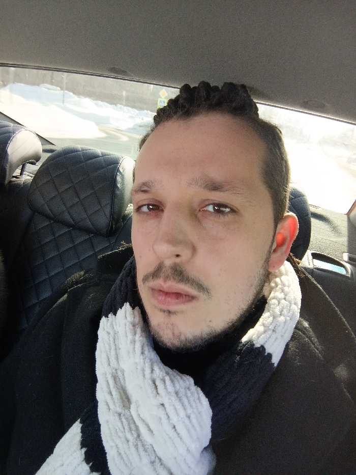 An adult male is depicted in a close-up, front-facing view, seated within the interior of a vehicle in Bessonovka, Russia. He has light skin, dark eyes with slight redness on the lower lids, a dark mustache, and a chin strap beard. His hair is styled with short, dark dreadlocks or braids on the crown and shorter sides. He is wearing a dark, high-neck garment under a dark coat, and a thick, textured scarf with alternating black and white horizontal stripes is wrapped around his neck. Behind him, black diamond-quilted leather headrests of the vehicle's seats are visible, along with a light grey ceiling liner. Through the vehicle's rear window, a brightly lit winter scene is observed, featuring snow-covered ground and a partial view of a yellow diamond-shaped road sign with a black symbol, located alongside what appears to be a paved road. The individual is positioned directly facing the camera.