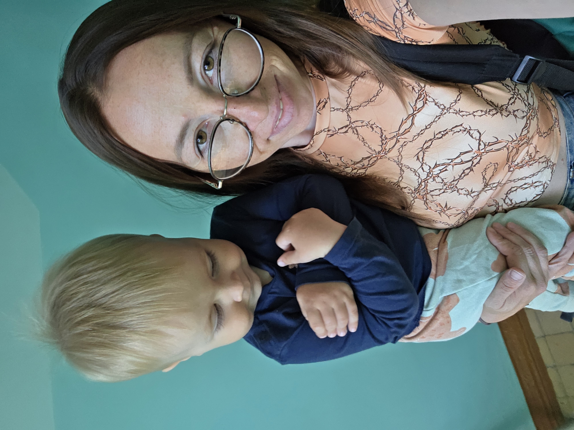 A woman wearing glasses and an orange patterned shirt holds a young child. The child, wearing a dark blue long-sleeved shirt and light blue patterned pants, is looking down with arms crossed. They are standing against a turquoise wall. The scene appears to be indoors, possibly in a home or public building in Chaikovsky, Russia.