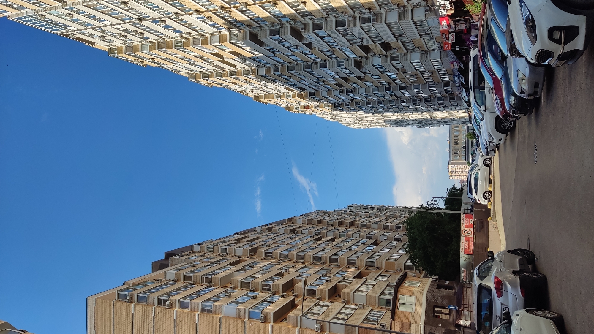 The image shows a street scene in the city of Krasnodar, Russia, with two tall, apartment buildings facing each other. The buildings are made of light-colored bricks and have many balconies and windows. Several cars are parked in the street between the buildings, which appears to be a residential area. The sky is blue with a few scattered clouds. A sign on the left building indicates a parking area. The overall impression is of a typical urban residential environment.