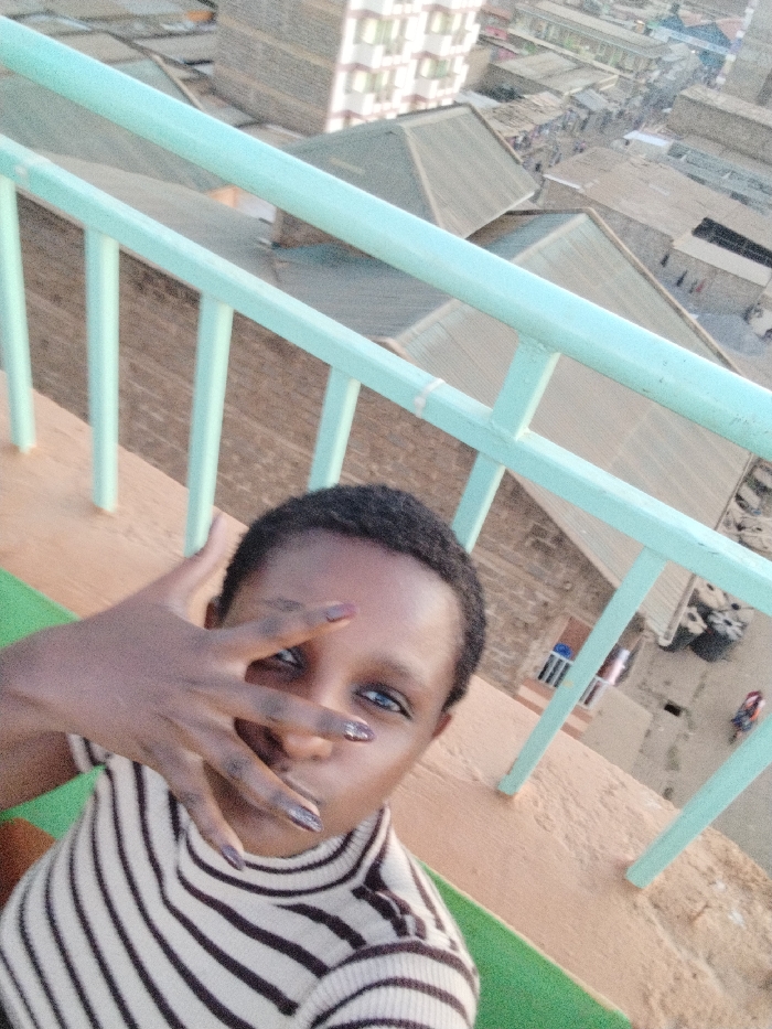 A woman wearing a striped sweater poses for a selfie with a hand over her face. She is positioned on a rooftop with a blue railing, overlooking a dense urban landscape. Buildings with corrugated iron roofs and a taller apartment block are visible in the background. The overall impression is one of a person taking a self-portrait from an elevated location.