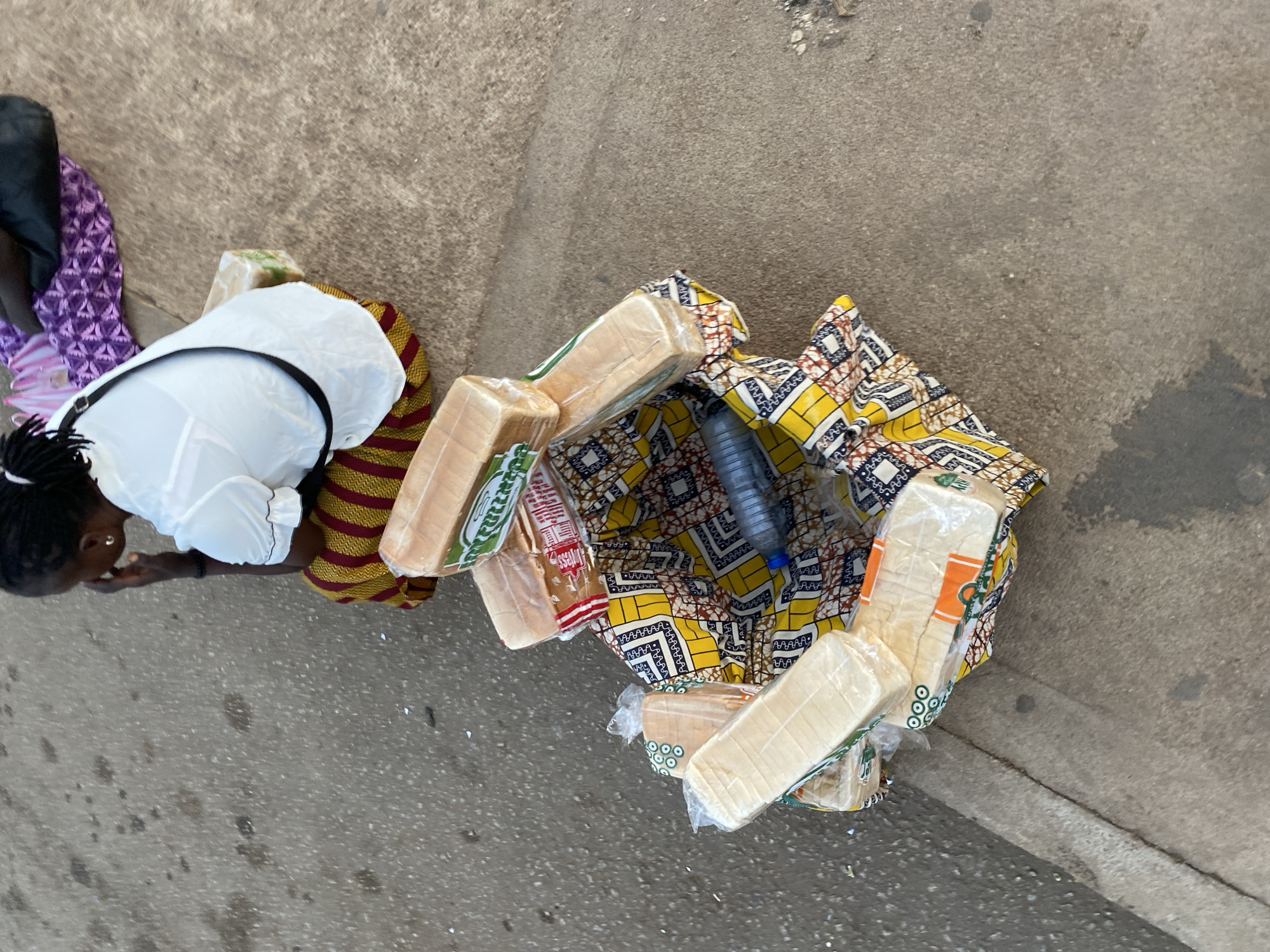 The image provides an overhead view of a scene in Bukuru, Nigeria, likely on a concrete or asphalt surface. A person, identified as a woman, is partially visible on the left side, appearing to kneel or bend down. She wears a white blouse with dark trim and a skirt or lower garment made of striped fabric in red and yellow/gold hues. Her hair is styled in dark braids or dreadlocks, and she has an earring in her visible ear.
Arranged on the ground to her right is a vibrant collection of items, prominently featuring multiple loaves of white bread. The bread loaves are individually wrapped in clear plastic packaging. Several loaves display colored labels; one clearly shows the word "Tess" in white lettering against a red background, while another has a green patterned label. These loaves are laid out on a large, intricately patterned piece of textile, primarily in yellow, blue, red, and white. A clear plastic water bottle with a blue cap is also visible among the bread and fabric.
The arrangement suggests a street vending setup or the transportation of goods, characteristic of a market environment. The lighting indicates it is daytime.