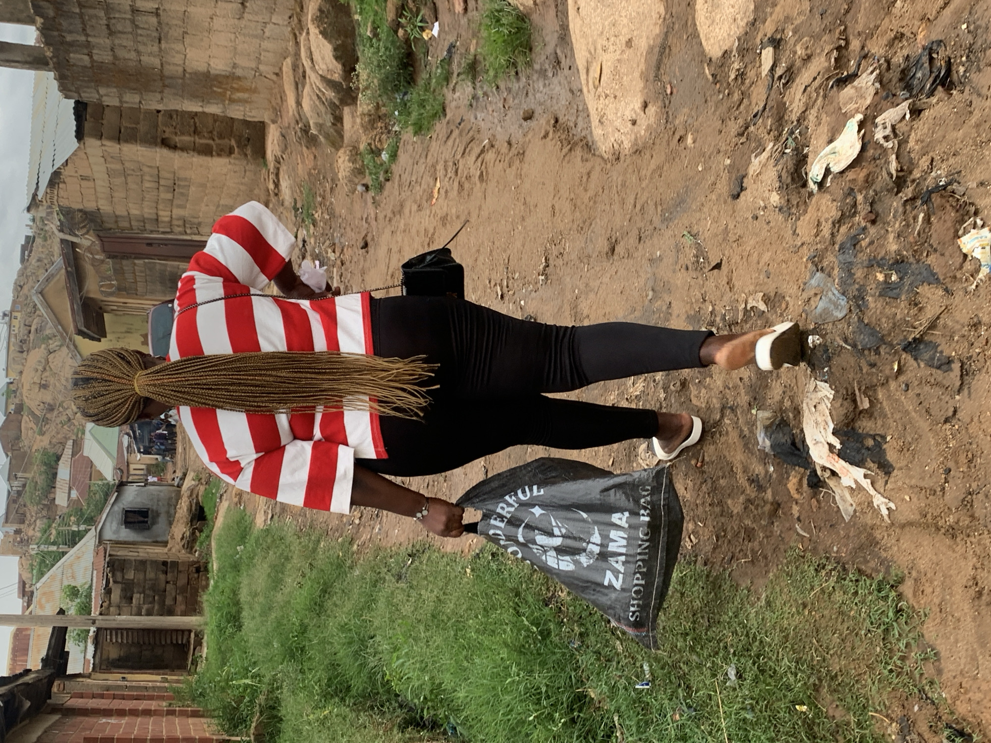 A woman walks on a dirt road in Jos, Nigeria. She wears a red and white striped shirt, black leggings, and white sneakers. She has long, braided hair and a black purse slung across her body. She carries a black shopping bag with "ZAMA SHOPPING BAG" printed on it. The road is muddy and littered with trash. Buildings and rocks are visible in the background. Grass is growing on either side of the path.
