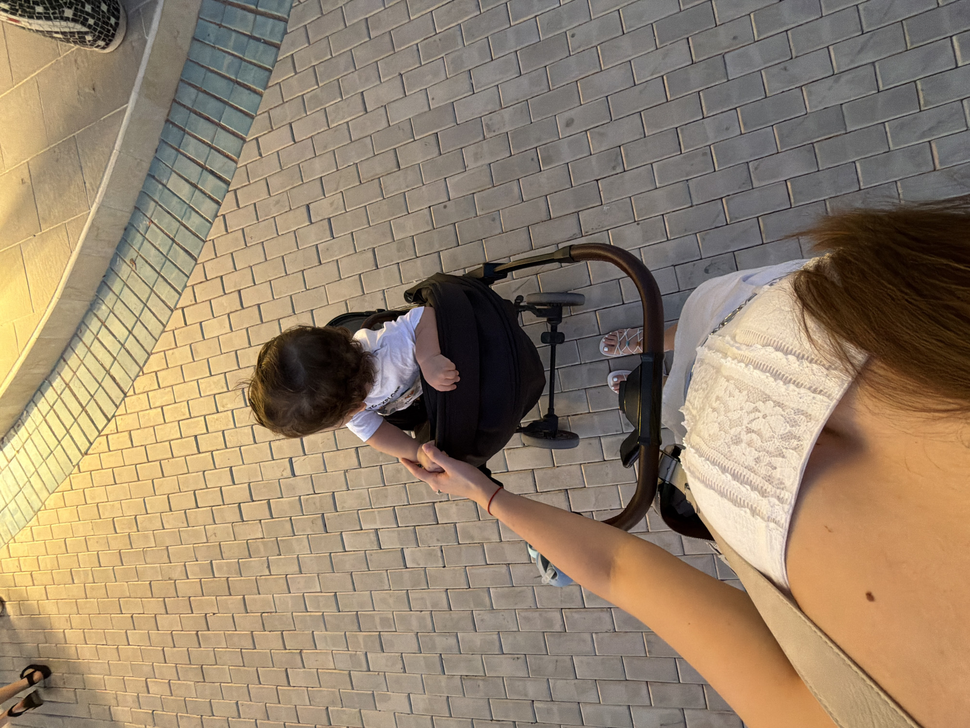 The image shows a high-angle, close-up view of a baby sitting in a stroller, with a person's arm reaching towards the baby. The baby has dark curly hair and is wearing a white t-shirt. The person's arm is slender and has a red bracelet on the wrist. The stroller is black with brown handles. The scene appears to be outdoors on a paved walkway made of light-colored rectangular bricks. In the background, there is a section of a curb with lighter-colored bricks and a blue-tiled section. There are also hints of other people's legs and feet in the upper left and upper right corners, wearing dark sandals and patterned shoes, respectively. The lighting suggests it is either early morning or late afternoon. The environment is consistent with an outdoor public space in Sharm El Sheikh, Egypt.
