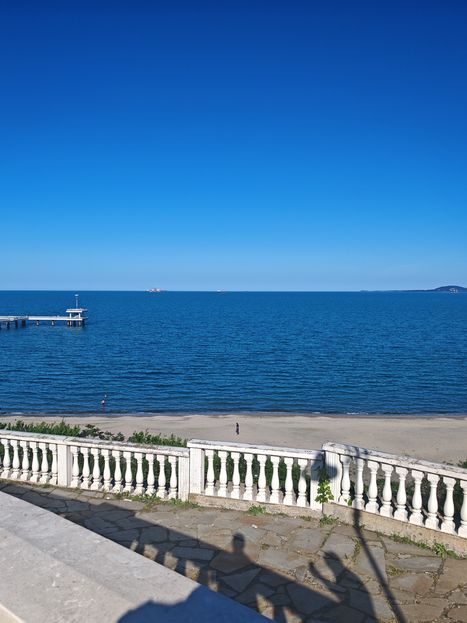 The image depicts a coastal scene in Burgas, Bulgaria, under a clear blue sky. The foreground features a white balustrade made of concrete, partially obscuring the view. The midground reveals a sandy beach with a few people, including a lone figure walking near the water's edge. The sea stretches to the horizon, marked by distant ships. To the left, a pier extends into the water, featuring a structure at its end. The water is a deep blue, reflecting the sky. The ground is covered with stone tiles. Shadows of people are cast on the ground.