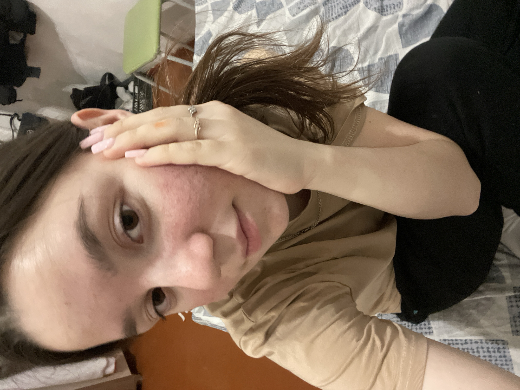A person is lying on a bed in an interior setting. The person is partially visible, wearing a tan-colored t-shirt and black bottoms. Their left hand is placed on their face, revealing a ring on one of their fingers and light pink nails. The bed is covered with a patterned blue and white sheet. In the background to the left, there's a glimpse of a light green object, possibly a chair, and other indistinct items. The image is a selfie style shot, taken at a close angle to the subject's face. There is no verifiable evidence to indicate the location in the city of Ольховка, Russia.