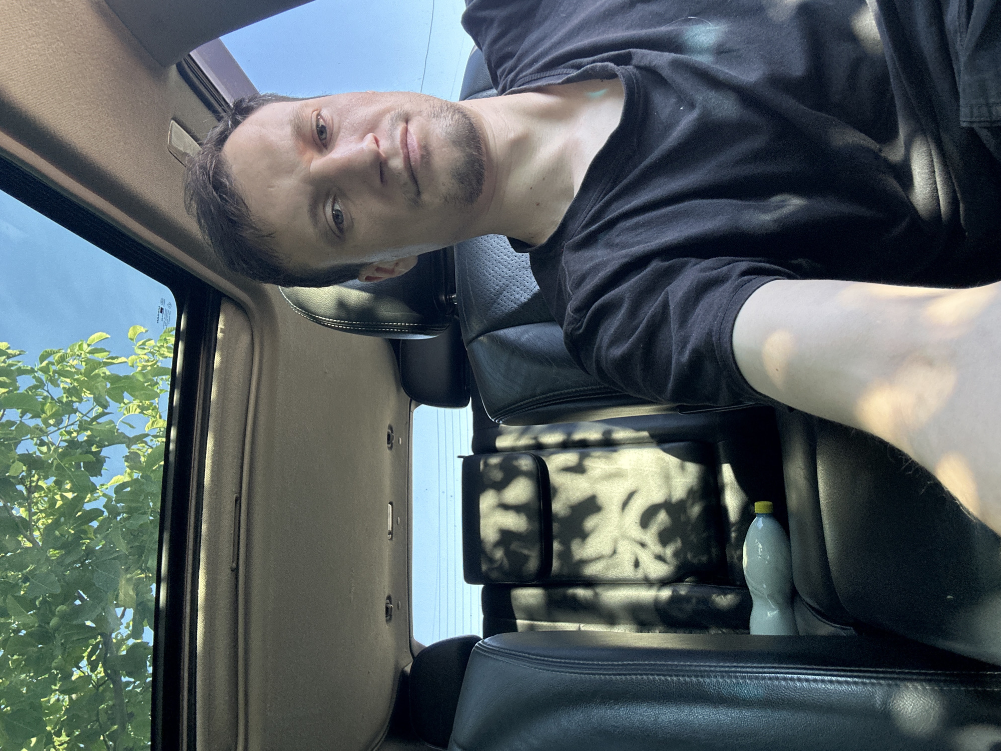 A man is sitting in the driver's seat of a car. He is wearing a black t-shirt. The car has a sunroof and the interior is dark leather. Sunlight filters through the trees, casting shadows on the interior of the car. A bottle of liquid is visible on the seat behind the driver. The scene appears to be a parking area or street in Shatava, Ukraine.