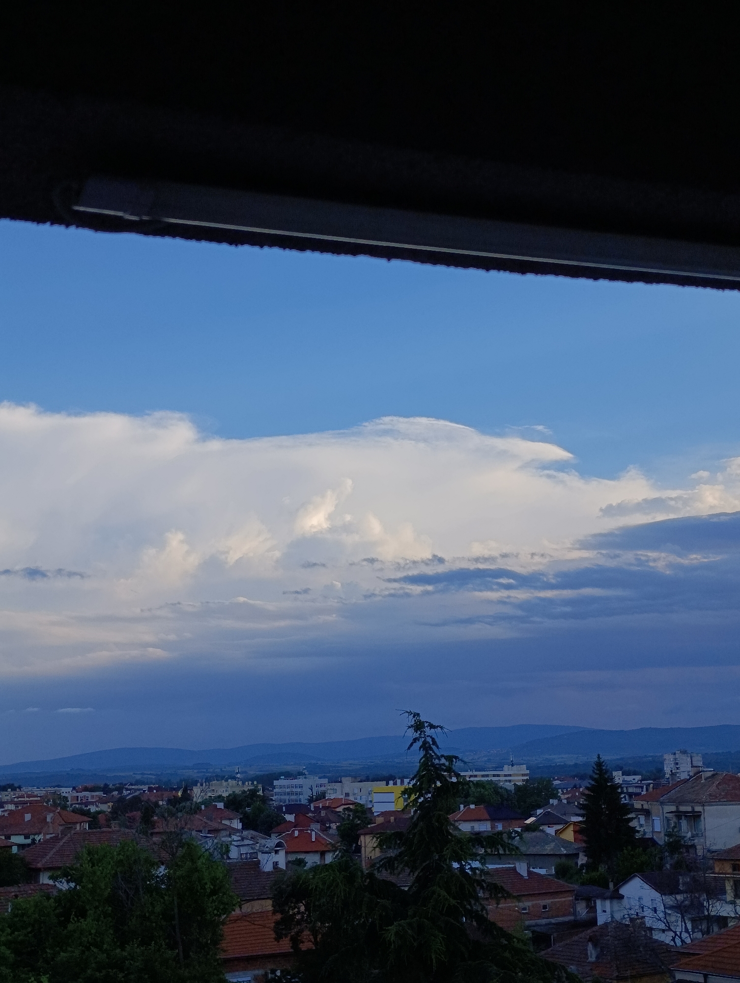 The image captures a cityscape in Svilengrad, Bulgaria, from an elevated perspective, likely through a window. The sky is predominantly blue, transitioning to darker tones towards the horizon, with large, white cumulus clouds dominating the upper part of the frame. The lower portion of the image reveals a densely packed residential area with red-tiled roofs and interspersed trees. In the distance, the silhouette of a mountain range is visible. A dark, blurred element, likely part of the window frame, occupies the top edge of the image.