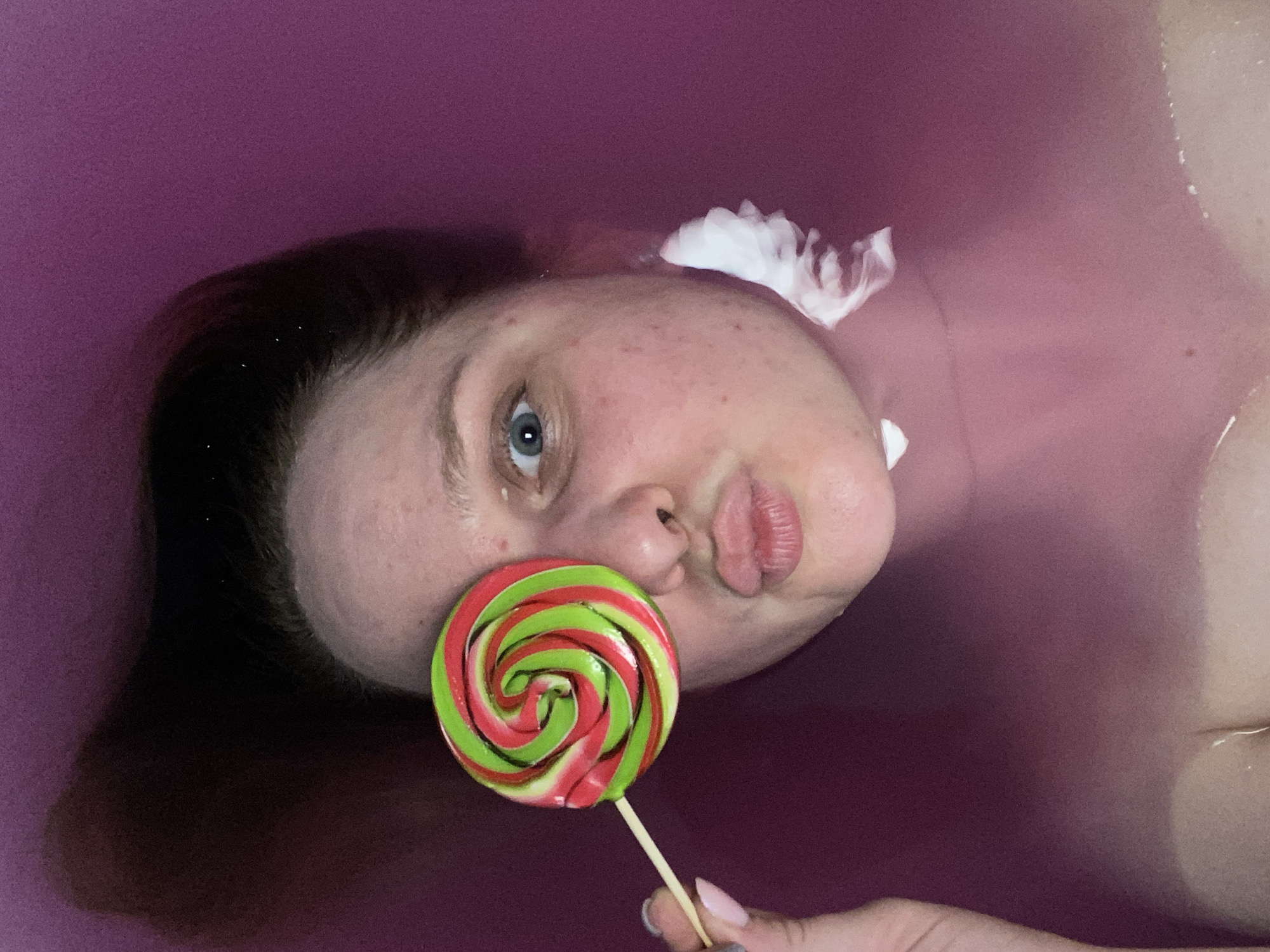 A close-up, overhead, indoor shot captures a person submerged in pink water, holding a red and green swirled lollipop to their right eye. The individual's face is turned towards the camera, with their lips puckered. Their hair is dark and appears wet, fanning out behind them in the water. White sudsy foam is visible on the right side of the person's face, near their ear. The background is a solid, dark pink color. The setting is identified as being in Chaikovsky, Russia.