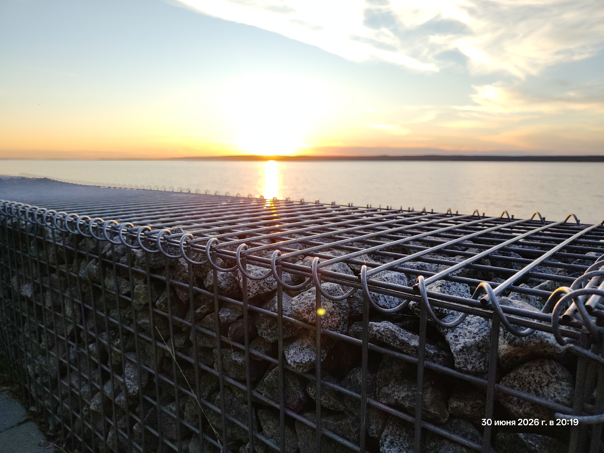 This image depicts a sunset over a calm body of water, likely a lake or the sea. In the foreground, a gabion wall, constructed from wire mesh filled with rocks, runs along the edge of the water. The setting sun casts a warm, golden glow across the sky and reflects on the water's surface, creating a serene and peaceful atmosphere. The sky transitions from a pale blue at the top to a vibrant orange and yellow near the horizon. Scattered, wispy clouds are visible, catching the light of the setting sun.
The gabion wall is the dominant object in the foreground, its wire mesh structure filled with rough, grey stones of varying sizes. The texture of the stones and the metal of the mesh are clearly visible. Sunlight glints off the metal rings that bind the mesh, adding a subtle sparkle to the scene. The water appears placid, with only slight ripples. The overall mood is one of tranquility and the quiet beauty of nature at the end of the day.
Visible text in the bottom right corner reads: "30 июня 2026 г. в 20:19", which translates to "June 30, 2026 at 20:19". This indicates the date and time the photograph was taken. No people are present in the image. The weather appears clear, with the sun shining brightly despite being low on the horizon. The location is not explicitly identified, but the presence of a large body of water and a gabion wall suggests a coastal or lakeside environment.