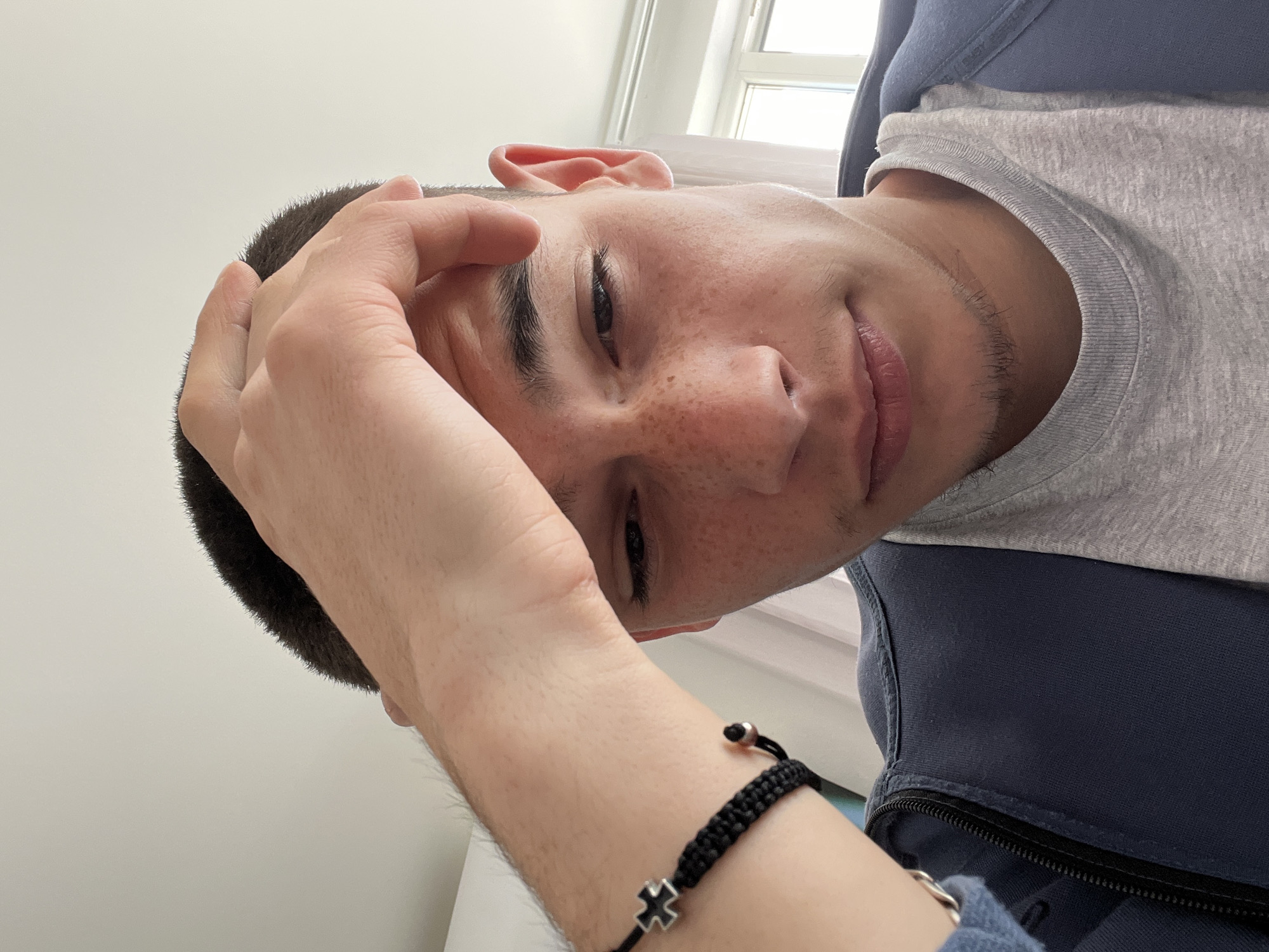 A young man with a short haircut, fair skin, and freckles is looking at the camera from a tilted angle. He is wearing a grey shirt and a dark blue hooded jacket. His left hand is raised to his forehead. He is wearing a black braided bracelet with a silver cross and bead. The background is a white wall with a window. The image location is not specified within the provided image.