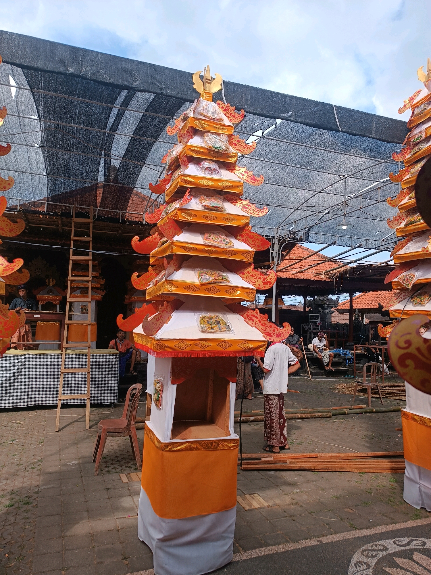 A man wearing a white shirt and a traditional sarong stands in front of a tall, tiered structure adorned with orange and gold fabric, suggesting a ceremonial or decorative purpose. The structure is topped with golden finials. The man appears to be facing away from the camera, with his hands resting on his hips. In the background, a black checkered cloth is draped over a structure, with a wooden ladder leaning against it. Several other people are visible in the background, some sitting and some standing, in what appears to be an outdoor setting. The environment is paved with stone tiles, and there is a large black mesh covering the area, supported by metal poles, creating a shaded space. Wooden planks and bamboo poles are scattered on the ground. The overall scene suggests a gathering or preparation for an event in Selemadeg, Indonesia, possibly a religious ceremony or festival, given the ornate decorations and traditional attire.