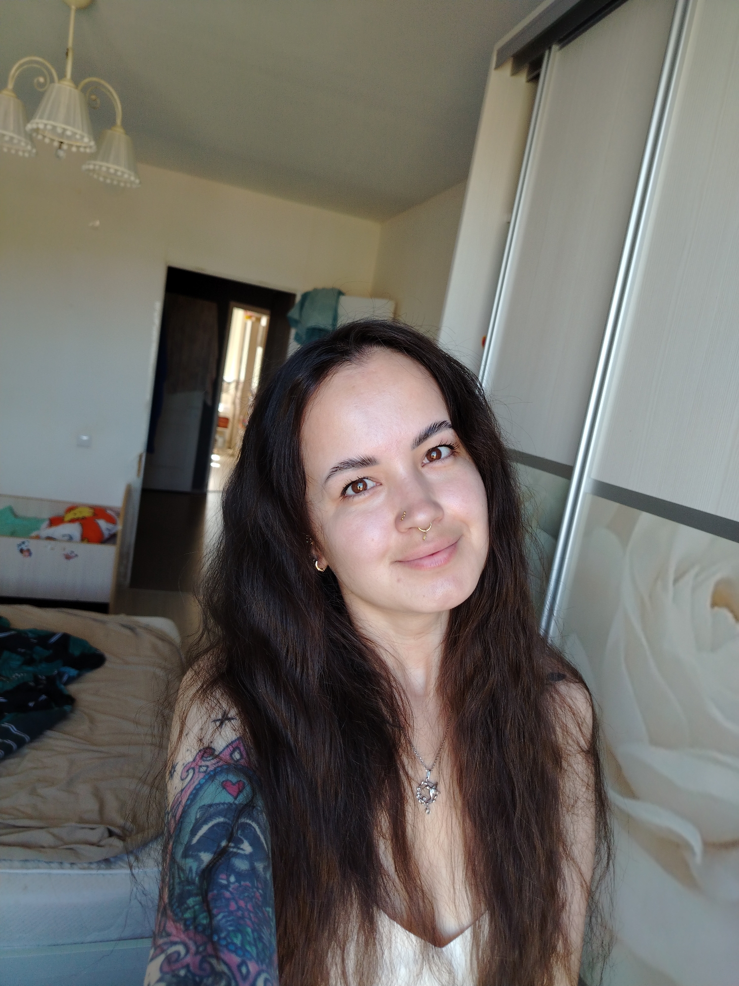 A woman with long, dark hair and a nose ring is the subject of a medium shot. She is wearing a necklace and has a tattoo on her left arm. She is positioned in an interior space, possibly a bedroom. In the background, there is a bed with a patterned blanket, a doorway leading to another room, and a large wardrobe with sliding doors, one of which features a floral pattern. The room appears to be illuminated by natural light. There is no indication of the location being in the city of Noviy, Russia.