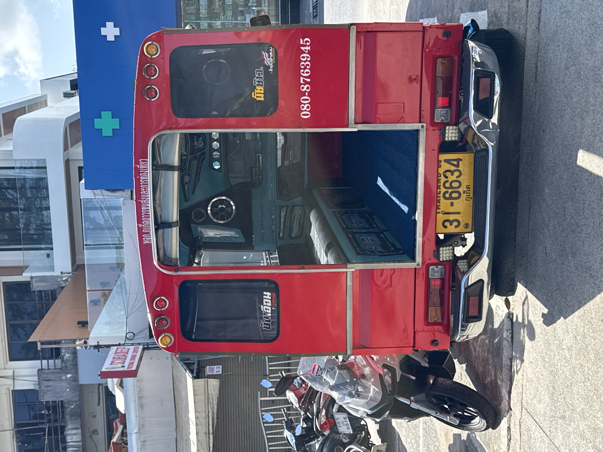 The image shows the rear of a red tuk-tuk parked on a street in Patong, Thailand. The tuk-tuk has a license plate that reads "31-6634" and "Phuket" below it. There are two motorcycles parked to the left of the tuk-tuk. In the background, there is a building with a blue facade and green and white cross symbols. The street is paved with concrete and there are shadows cast by the objects in the scene. There are no people visible in the image.