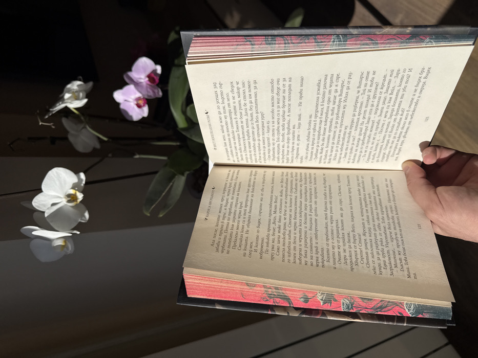 A human hand is captured indoors, holding an open book written in Bulgarian Cyrillic. The book is open to pages 122 and 123. The left page displays the name "КАРИСА БРОУДЪНТ" (Carissa Brouden), and the right page shows "ТЪЙ ИЗСТЪПЕНИ РОЗИ" (So Steeped Roses). The book's visible spine and fore-edge feature a decorative pattern with red and black stylized motifs. In the background, an out-of-focus orchid plant is visible, featuring several white blossoms in the upper left and fainter pink blossoms further to the right. Strong natural light, originating from the left, illuminates the book and the prominent white orchids, creating a high-contrast scene with dark background areas. The setting is an indoor residential or private space in Sofia, Bulgaria.
