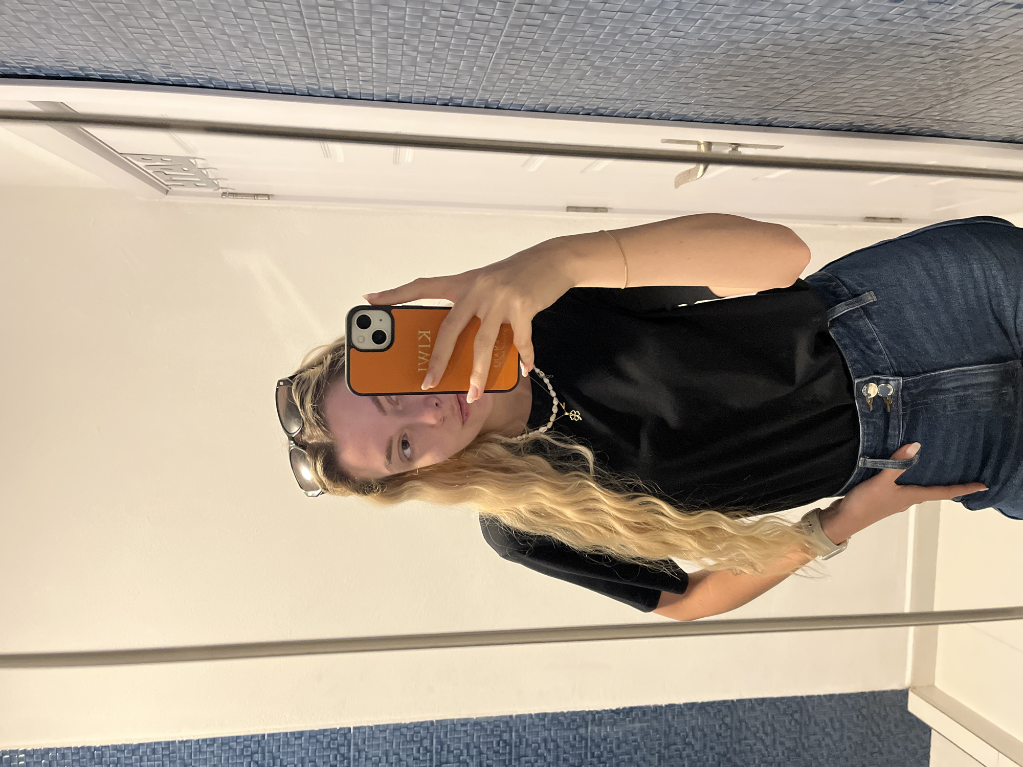 A young woman is depicted taking a mirror selfie, with the image currently rotated 90 degrees counter-clockwise. She has long, wavy blonde hair that falls over her shoulders. Her gaze is directed towards the phone/mirror with a neutral expression.
She is wearing a black short-sleeved top and blue jeans. Accessories include sunglasses perched on top of her head, a pearl necklace with a gold-colored clover pendant, a simple gold-colored bracelet on her right wrist, and a light-colored watch on her left wrist.
Her right arm is raised, holding an orange smartphone with a black camera module. The phone case clearly displays the text "KIMI" and "SAVI" (appearing mirrored as "IMIK" and "IVAS" in the image). Her left hand is resting near her hip/waistband.
The background, visible in the mirror's reflection, shows white walls with sections of small blue square tiles along the bottom and top edges, suggesting an indoor setting like a bathroom or a hallway. A white door frame and a horizontal metal bar, possibly part of the mirror's frame or room architecture, are also visible. No specific time of day or weather conditions can be discerned.