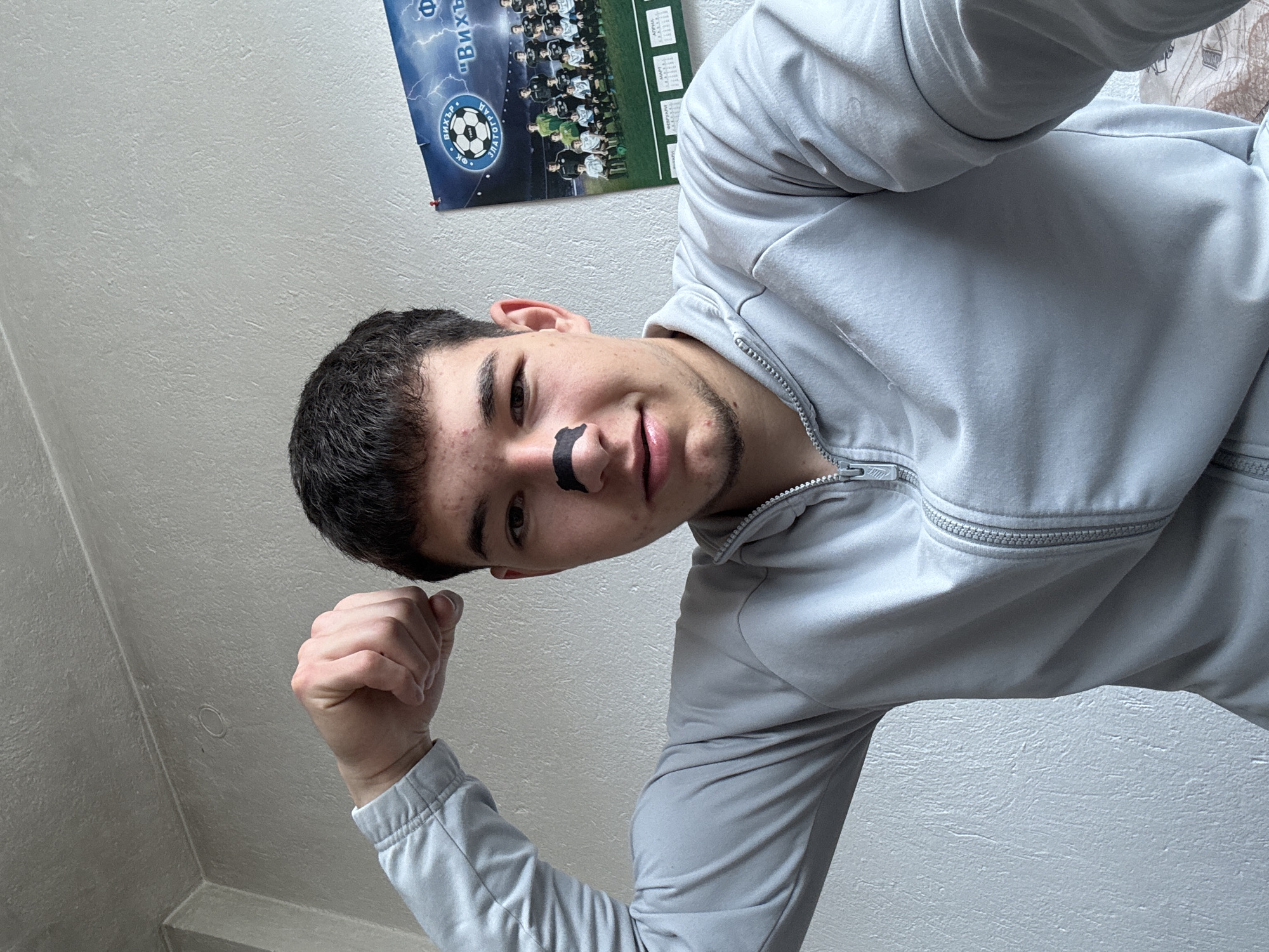 A young man stands indoors, wearing a gray zip-up jacket, and flexing his fist. He has short, dark hair, a small amount of facial hair, and a black adhesive strip on his nose. Behind him, attached to a light-colored wall, hangs a poster displaying a soccer team. The poster features the text "Вихър" and a soccer ball image. The setting is presumed to be an indoor space, possibly a residence, in Zlatograd, Bulgaria, judging by the Cyrillic text on the poster.