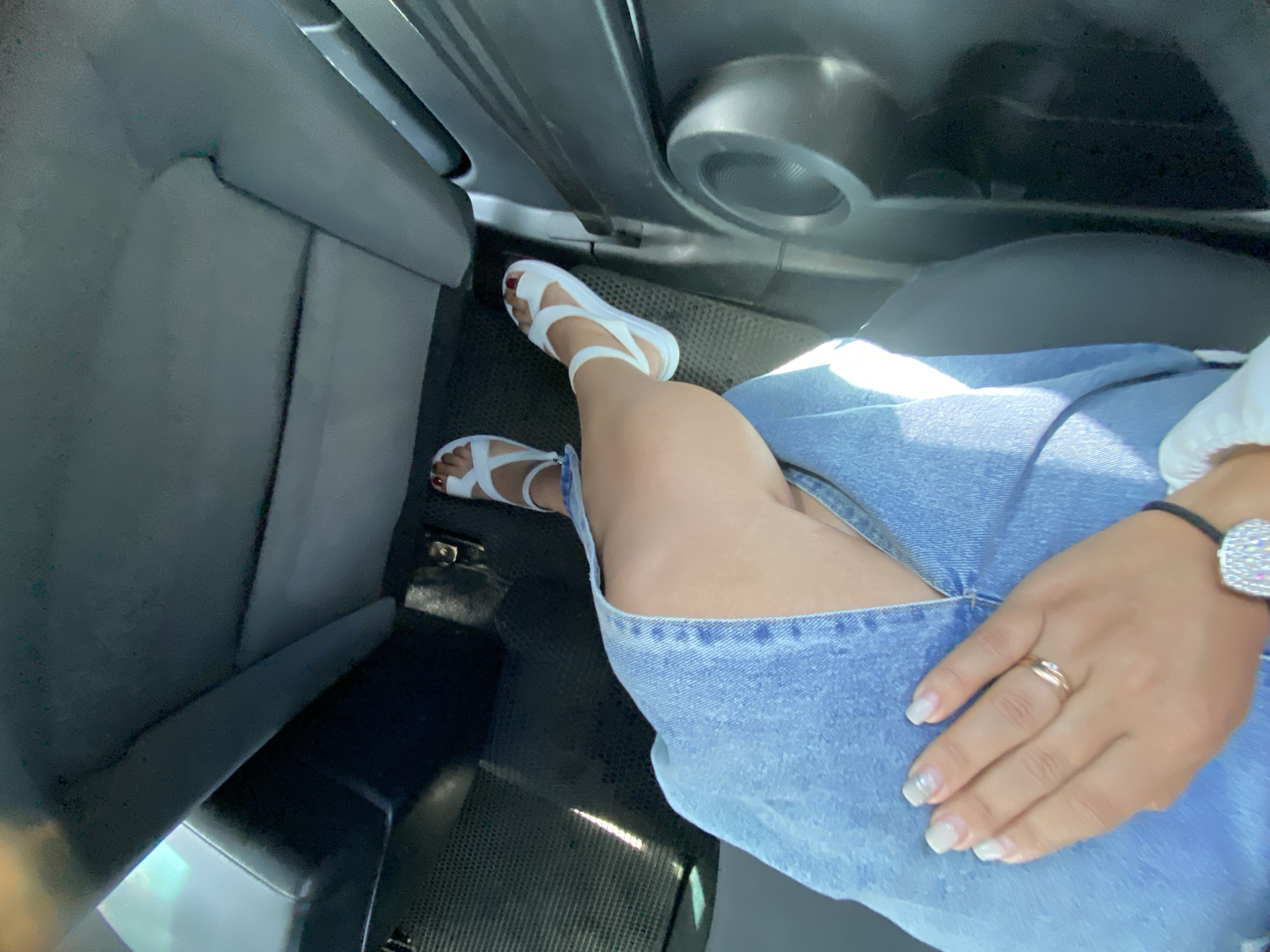 The image shows a person's legs and hands inside a car. The person is wearing white sandals and a denim skirt. Their hand is resting on their leg, and they are wearing a ring on their finger. The car's interior is visible, including the seats and a speaker. The location is the city of Tyulyugul, Russia.