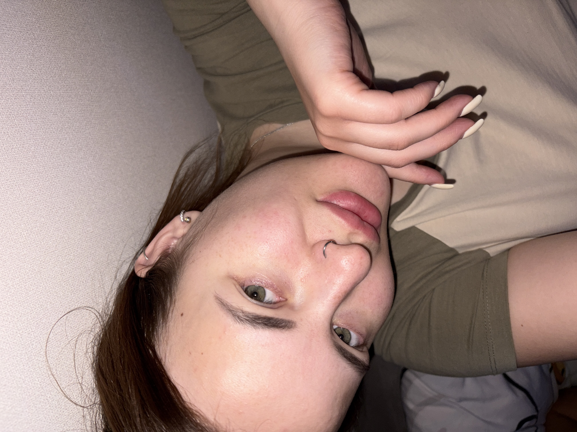 A close-up, rotated image shows a light-skinned woman lying down. She has light brown hair, fair skin, and green eyes. She has a silver earring in her left ear and a silver nose ring in her left nostril. Her fingernails are long and painted a light color. She is wearing a two-toned t-shirt with one side olive green and the other side beige. The background is a light-colored wall. The location and city are Unknown, Unknown.