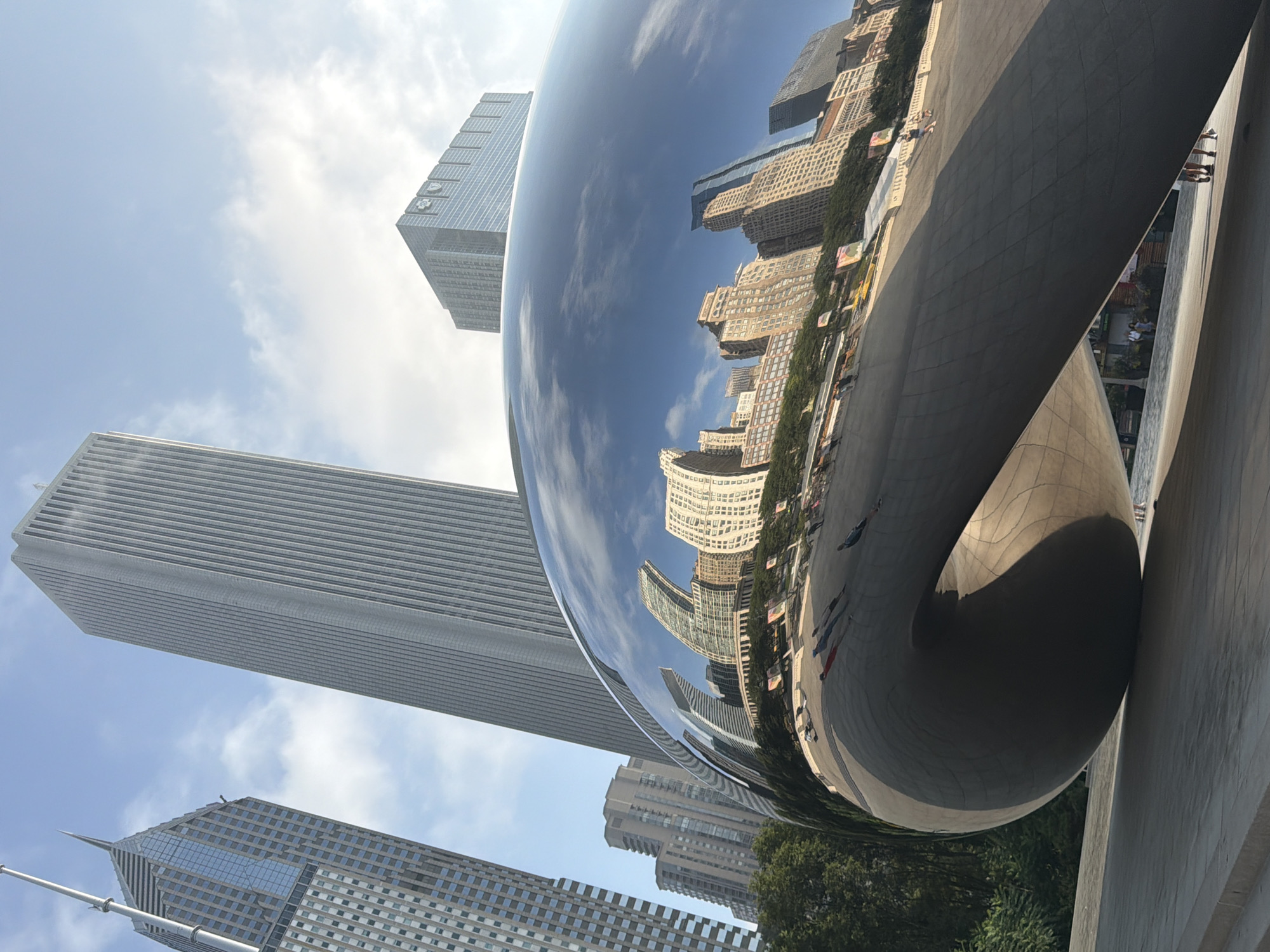 The photograph captures a dynamic perspective of Cloud Gate, a public sculpture located in Millennium Park, Chicago, Illinois, United States. The sculpture, commonly known as "The Bean," is a highly reflective, metallic surface that mirrors the surrounding cityscape and sky. In the foreground, the curved, bean-shaped form of the sculpture dominates the lower half of the frame, with its polished surface reflecting the buildings and clouds.
The background features several prominent Chicago skyscrapers, including a tall, slender building with a spire on the left, and a much wider, vertically ribbed skyscraper to its right. Further into the background, another modern glass-faced building with a distinct square crown is visible.
The reflection on the sculpture's surface shows a cluster of mid-rise buildings with intricate facades, their details warped and stretched by the convex form. The sky above is a bright blue, dotted with wispy white clouds, which are also reflected in the sculpture.
Several people are visible in the distance, scattered across the plaza and on the steps leading up to the sculpture. They appear as small, silhouetted figures, some walking and some standing, their presence adding a sense of scale to the monumental artwork and urban environment. The plaza surrounding the sculpture is paved with concrete and has a network of subtle grid lines. Lush green trees are visible along the edge of the park, providing a contrast to the urban architecture. The lighting suggests it is daytime, with bright sunlight casting shadows and highlighting the reflective surfaces.