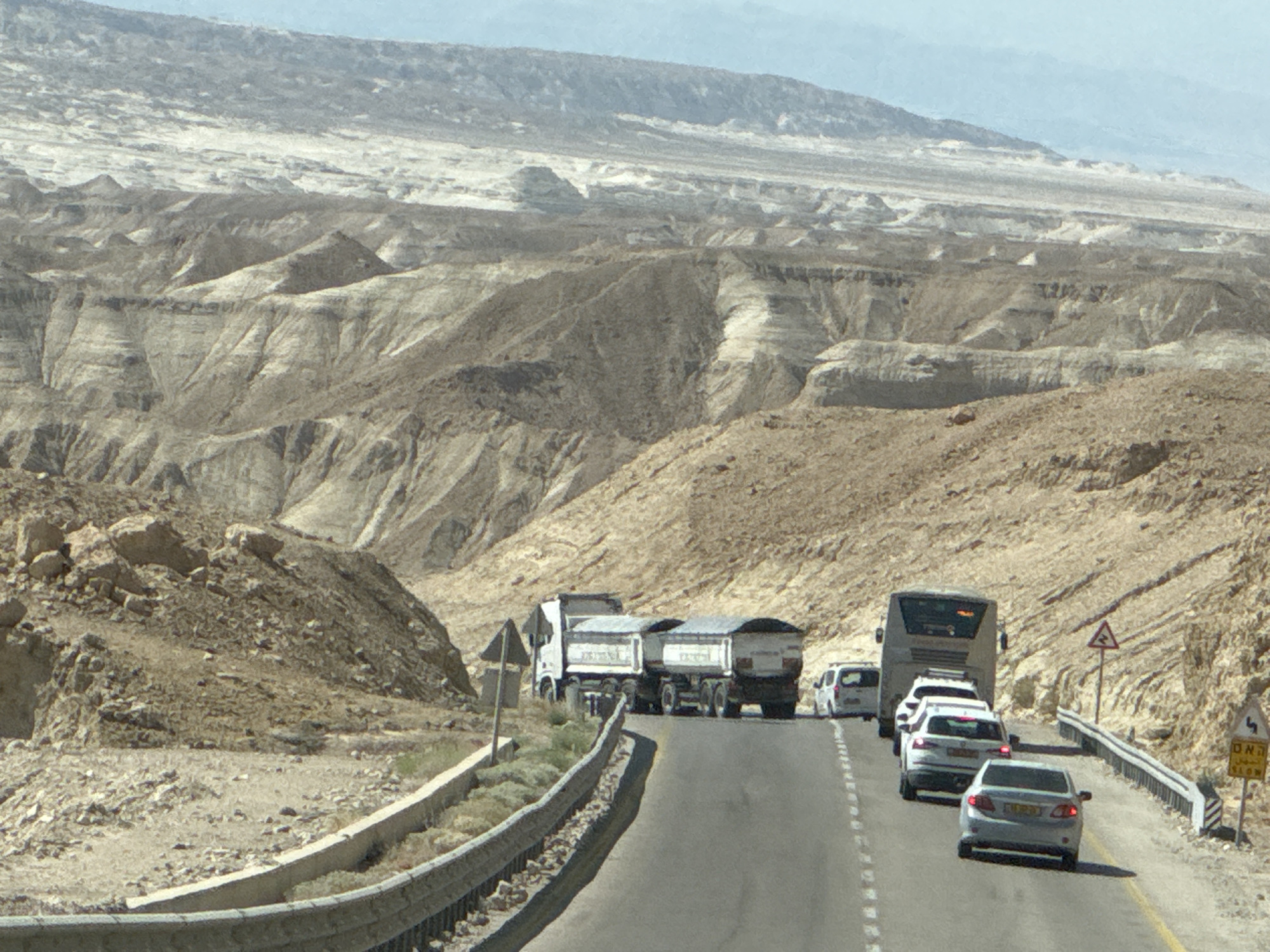 In the Tamar Regional Council, Israel, a convoy of vehicles travels along a winding asphalt road through a rugged, arid desert landscape characterized by eroded hills and rocky terrain. The road curves to the right, with a concrete barrier and guardrail lining the left side. In the foreground, a light-colored sedan is visible, followed by a white SUV and a grey sedan. Further ahead, a large tour bus leads a line of vehicles, including a white van. On the right side of the road, a yellow traffic sign with an arrow indicating a sharp curve and a warning of a hazard is posted. The background is dominated by the stark beauty of the desert mountains under a clear, pale blue sky. The scene suggests a journey through a remote and scenic part of Israel's desert region.