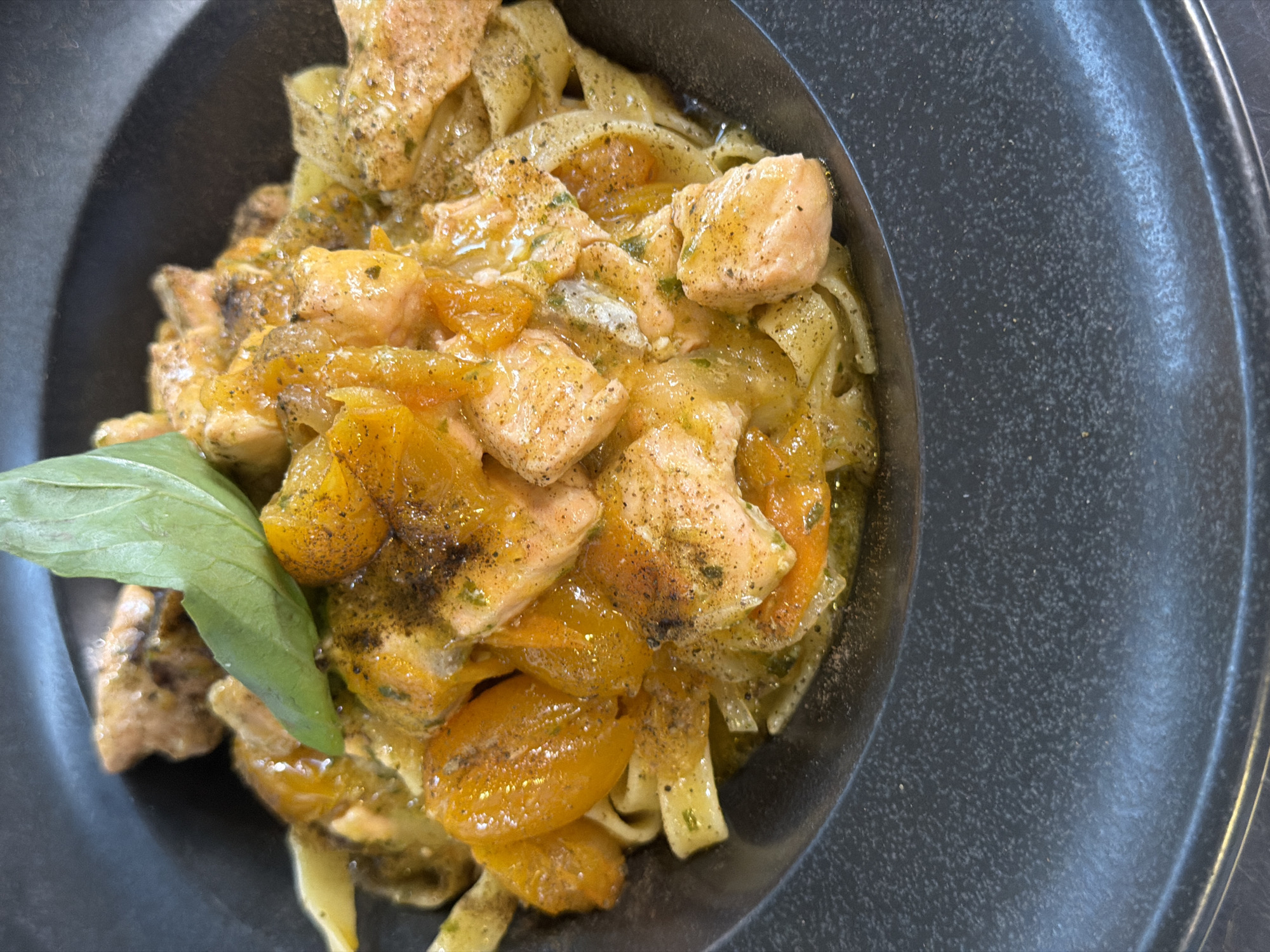 A close-up, top-down shot shows a dish of tagliatelle pasta with salmon and what appear to be cherry tomatoes in a shallow, dark gray, textured bowl. The pasta is coated in a light, yellowish sauce, with visible flecks of herbs and black pepper. The salmon is cut into bite-sized pieces and is mixed throughout the pasta. A single, large, green basil leaf rests on top of the dish. The lighting suggests an indoor setting, possibly a restaurant in Palmanova, Spain. There are no people visible in the image.