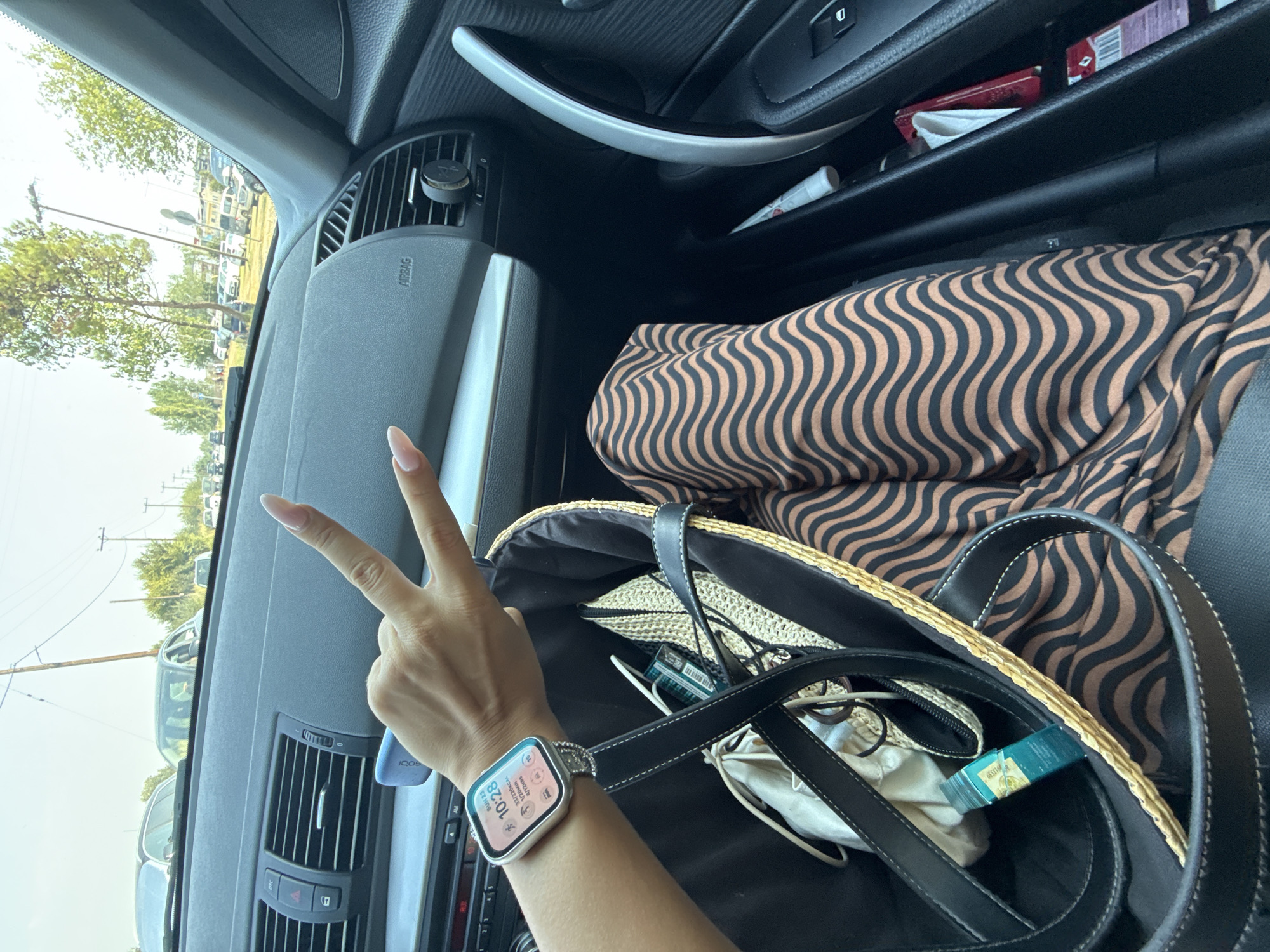 A first-person perspective shot taken from inside a car, looking forward. The driver, whose hand is visible making a peace sign, is wearing a smartwatch displaying the time as 10:28. Beside her, a straw-like bag contains a white pouch, a green bottle, and a woven hat. The driver is wearing a brown and black striped maxi dress. The car's interior, including the dashboard and air vents, is visible. Outside the car, a road with cars and trees can be seen, suggesting an outdoor, possibly suburban or rural setting.
