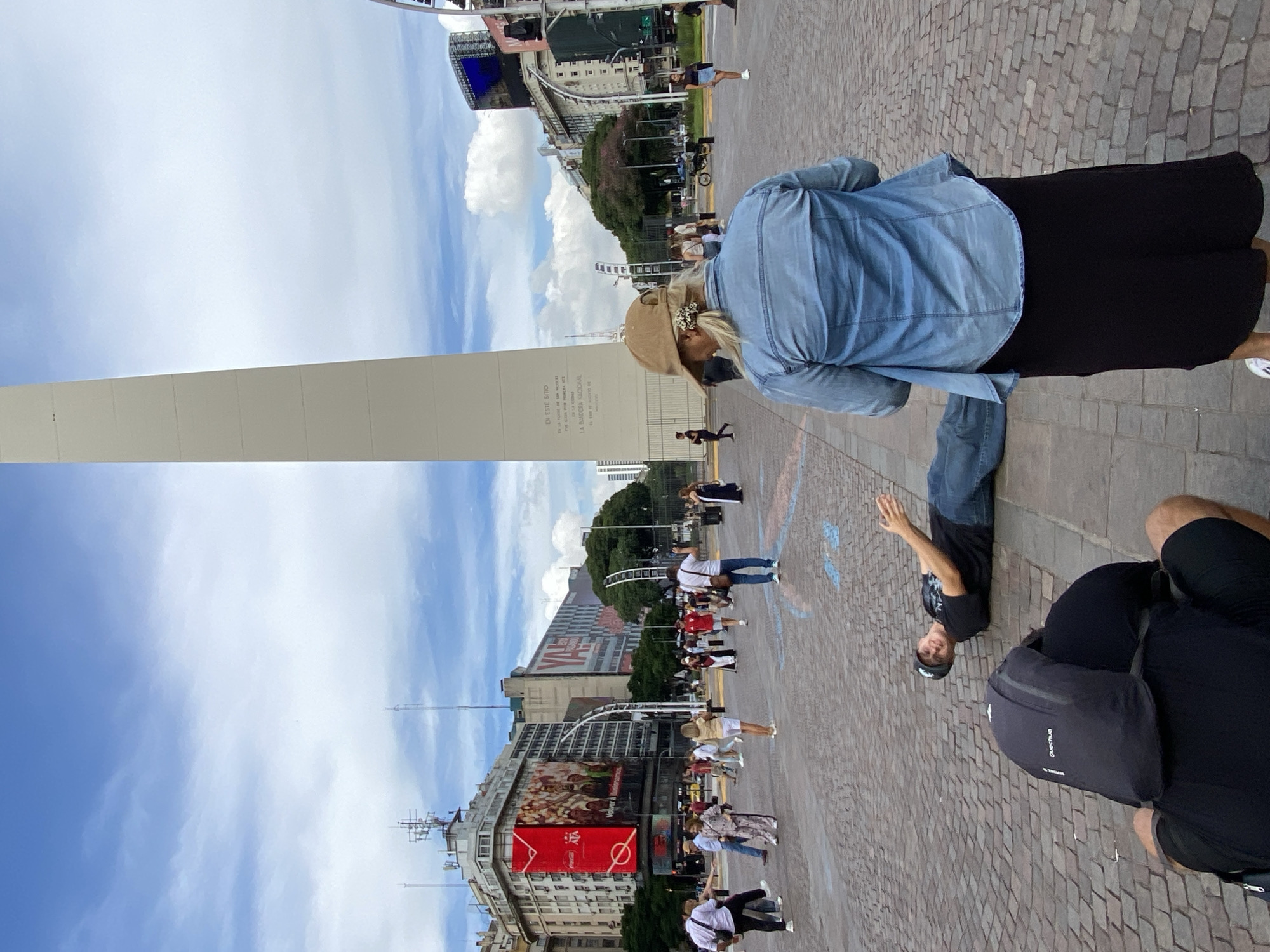 The image is a street scene in Buenos Aires, Argentina, featuring the Obelisco prominently in the background. It appears to be a busy day with many people walking around. There are buildings lining the street with advertisements displayed. In the foreground, there are people walking on the pavement. One person is wearing a denim jacket, while another is wearing a black backpack. The sky is partly cloudy.