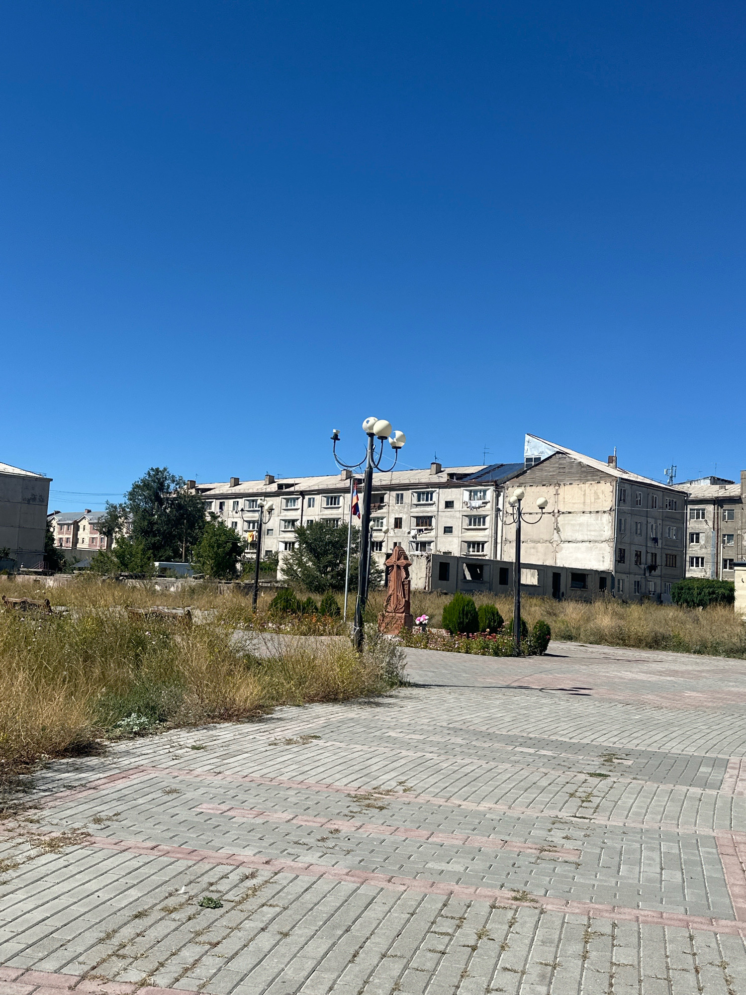 The image depicts a public square in the city of Gyumri, Armenia, with a clear blue sky overhead. In the foreground, a paved area made of grey and reddish-pink interlocking bricks is visible, with some patches of sparse grass growing between the stones. To the left, tall, dry grass grows in an unkempt area. In the mid-ground, a street lamp with three globes stands prominently. Near the lamp, a statue of a person, possibly religious in nature, is located. Behind the statue, a small flowerbed with green bushes and red and pink flowers adds a touch of color. Further in the background, a row of multi-story apartment buildings, characteristic of Soviet-era architecture, stretches across the scene. Some of these buildings appear to be in a state of disrepair, with exposed concrete and incomplete facades on one structure. Trees are scattered between the buildings and the open space. The overall impression is one of an urban environment that has seen better days, with a mix of residential buildings and public spaces that are not fully maintained.