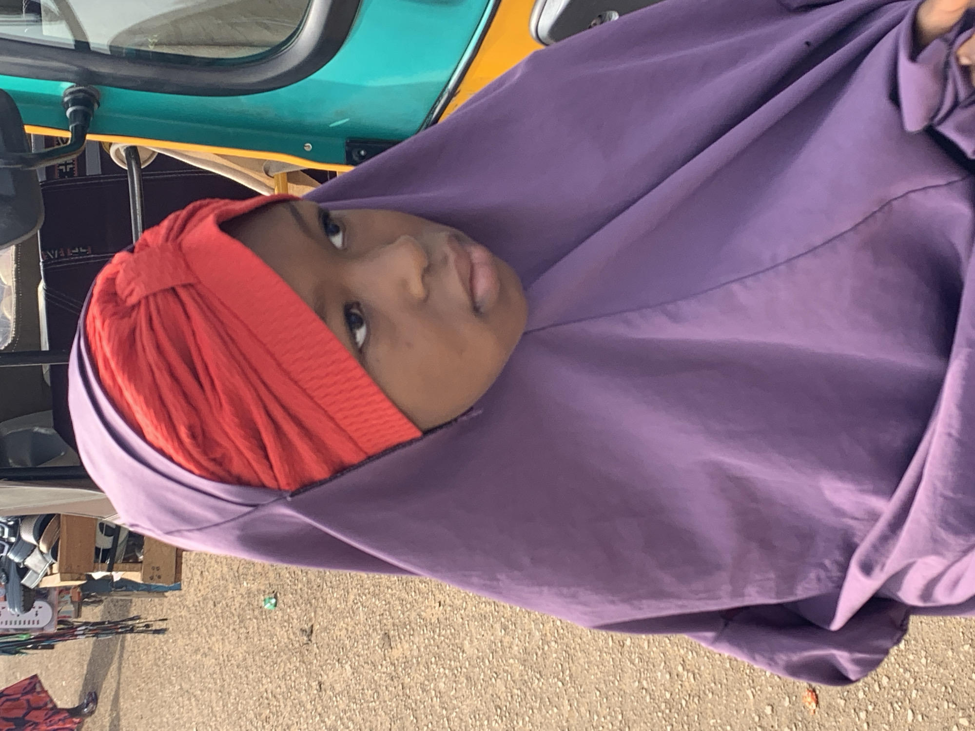 A young person wearing a red turban and a purple hijab is looking to the right. They are standing on a dirt road next to a turquoise and yellow auto-rickshaw. In the background, there is a market stall with various items, including shoes. The scene appears to be in a city.