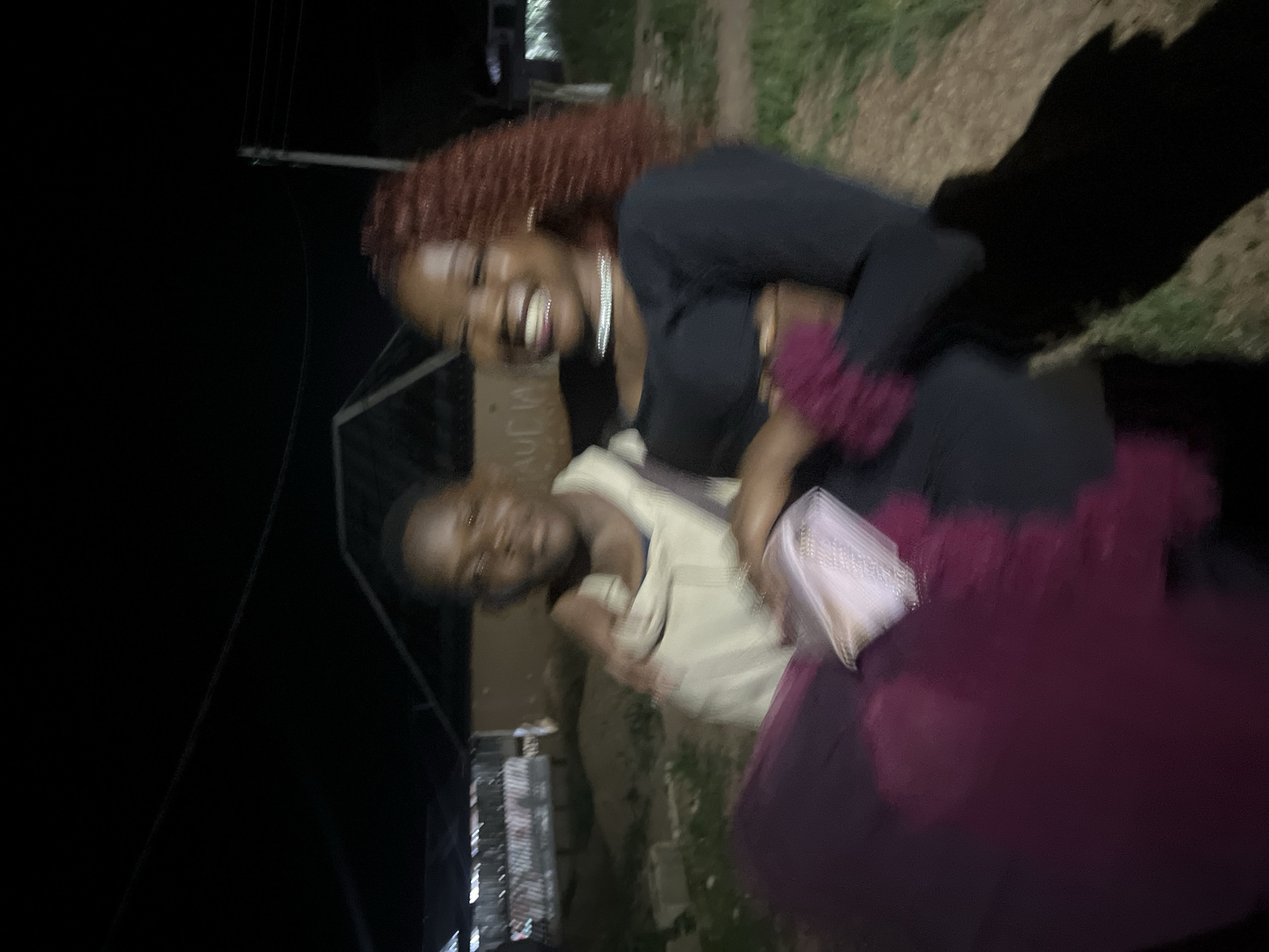 The image depicts two individuals, a younger person being held by an older individual, outdoors at night. The younger person, with dark skin and hair, wears a light-colored hooded garment. The older individual, with reddish-brown curly hair and dark skin, smiles broadly and wears a black long-sleeved shirt and a necklace. They are positioned outdoors, with a dark, indistinct background suggesting a rural or semi-urban setting. There are some structures in the background, possibly buildings or shelters, illuminated by faint light. The ground appears to be dirt and grass. The lighting is dim, casting shadows and creating a slightly blurred effect. The scene is consistent with a casual, nighttime gathering in a residential area within Durumi, Nigeria.