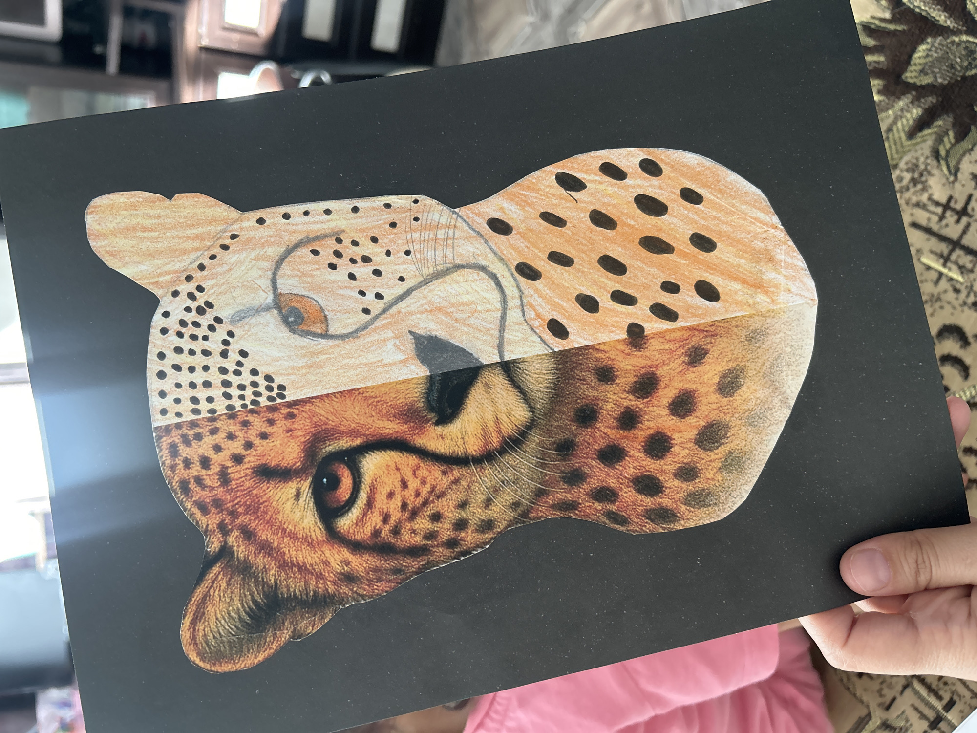A black rectangular piece of paper with a drawing/collage of a cheetah's head is being held up. The top half of the cheetah's head is a simple drawing with orange and black dots, while the bottom half is a more realistic rendering with detailed shading and coloring. The background behind the paper is blurry and appears to show furniture and part of a person wearing a pink shirt. The image was not taken in Uray, Russia, as the location is not discernible from the image itself.
