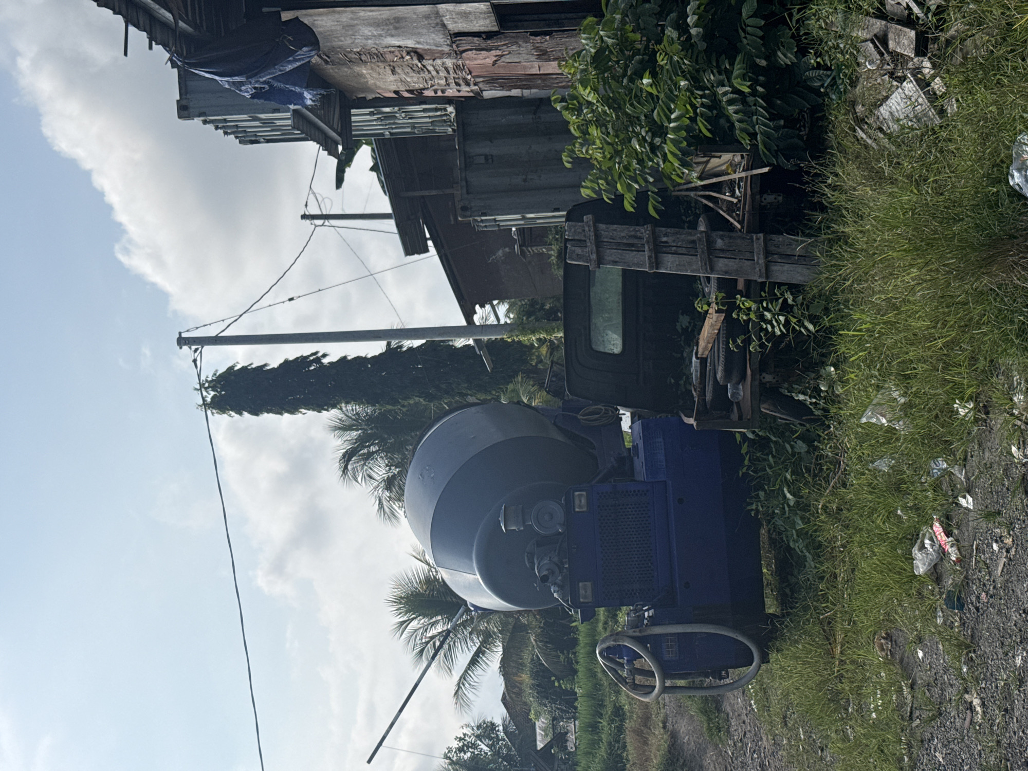 The image depicts an outdoor scene in Tampaksiring, Indonesia, characterized by a blue concrete mixer in the foreground. The mixer is situated on a gravelly path, surrounded by overgrown grass and scattered litter. Behind the mixer, a dark-colored truck bed is visible, along with various construction-related debris and wooden structures. Further in the background, palm trees and other vegetation suggest a tropical or subtropical environment. Several buildings, appearing somewhat dilapidated and constructed from corrugated metal and brick, line the right side of the frame, with power lines and utility poles visible overhead. The sky is partly cloudy. There are no people visible in the image.