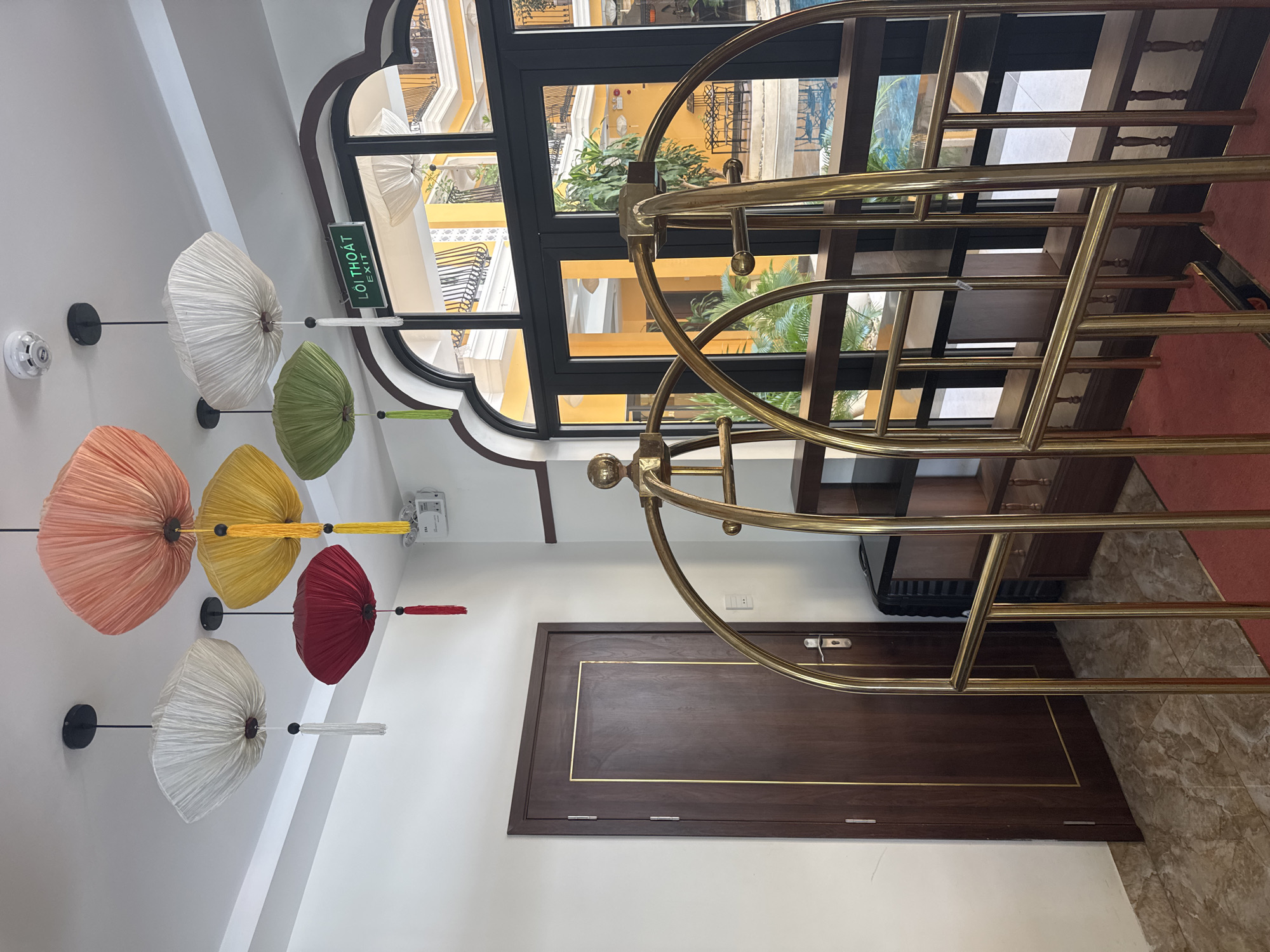The scene is an indoor shot of what appears to be the interior of a hotel or similar establishment in Đà Nẵng, Vietnam, based on the "Lối Thoát EXIT" sign. A luggage cart made of polished brass is positioned to the right, partially obscuring a dark wooden door with gold trim. To the left, a series of colorful paper umbrellas are attached to the ceiling as decorative elements, ranging in color from white to peach, yellow, green, and red. Above the luggage cart, a large, arched window reveals an exterior view of a building with yellow walls and black metal balconies. The overall lighting is bright and the environment suggests a modern, well-maintained interior.