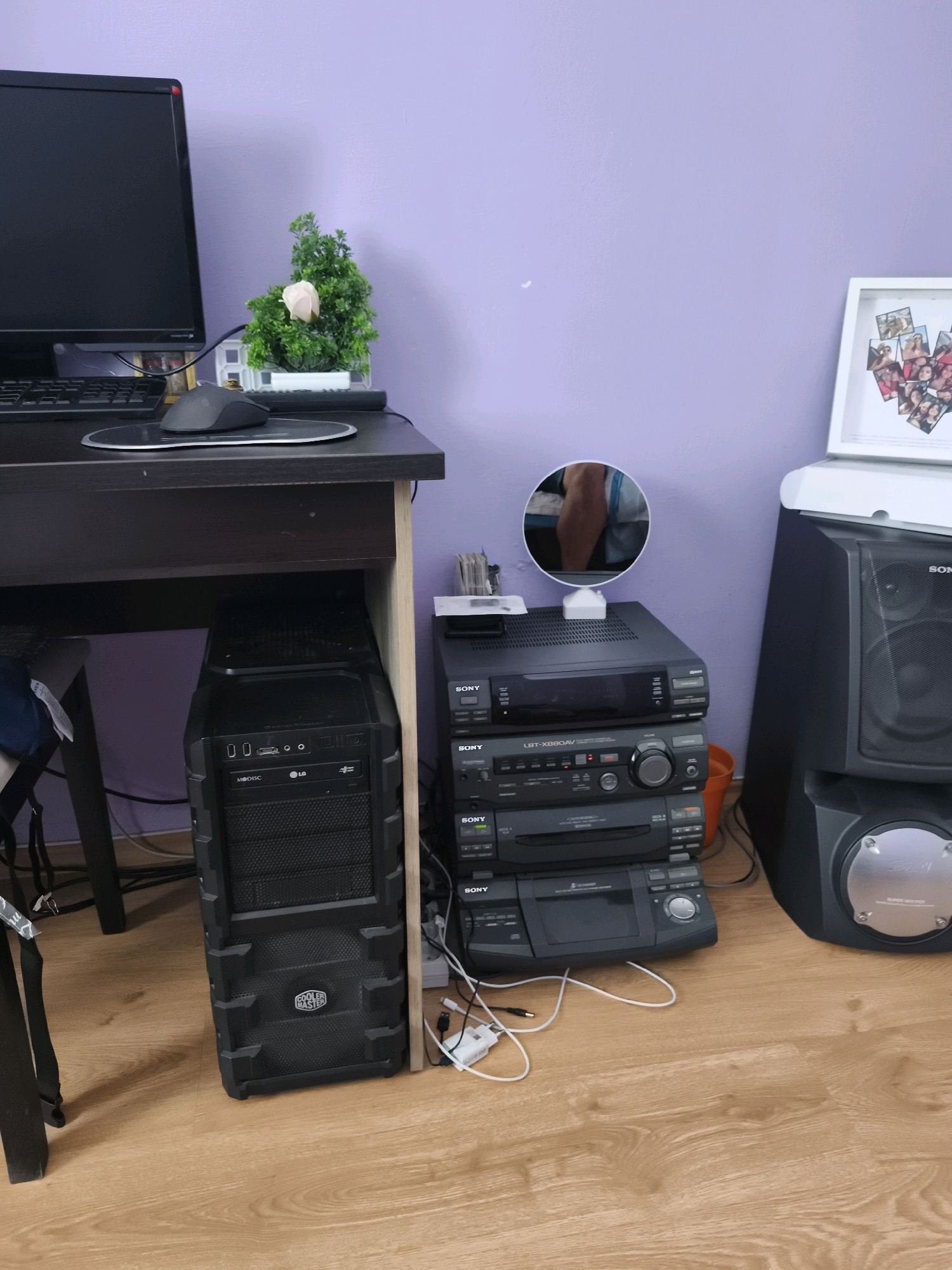 An indoor shot shows a wooden floor and a purple wall in a room. On the left, a black computer monitor is on a dark brown desk with a black mouse. Beneath the desk, a black computer tower is visible, with the "Cooler Master" logo on its front. To the right of the computer tower, a stack of three black Sony audio components are on the floor, with an orange bucket behind them. Further to the right, a large black speaker is positioned in the corner, topped with a white framed photo collage. A small potted plant with green leaves and a pink flower sits on the desk to the right of the monitor. A round mirror with a white base is placed on top of the uppermost Sony component, reflecting a person's forearm and hand.