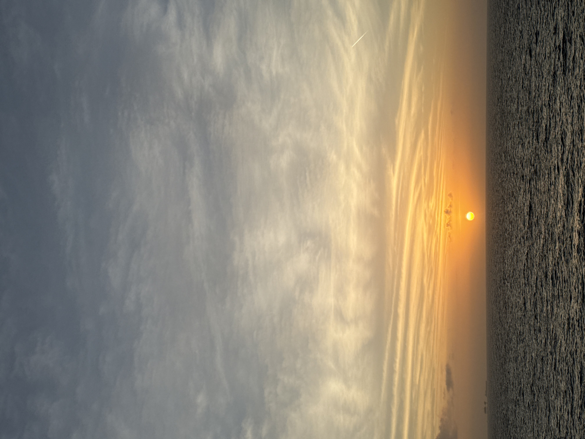 A landscape view of a horizon during sunset. The sky is filled with clouds in the left portion and streaks of golden, orange light in the right. The sun is a bright sphere near the horizon line. A dark body of water or textured land stretches from the horizon towards the bottom of the image. A contrail from an airplane is faintly visible in the sky. The location is Unknown, Unknown.