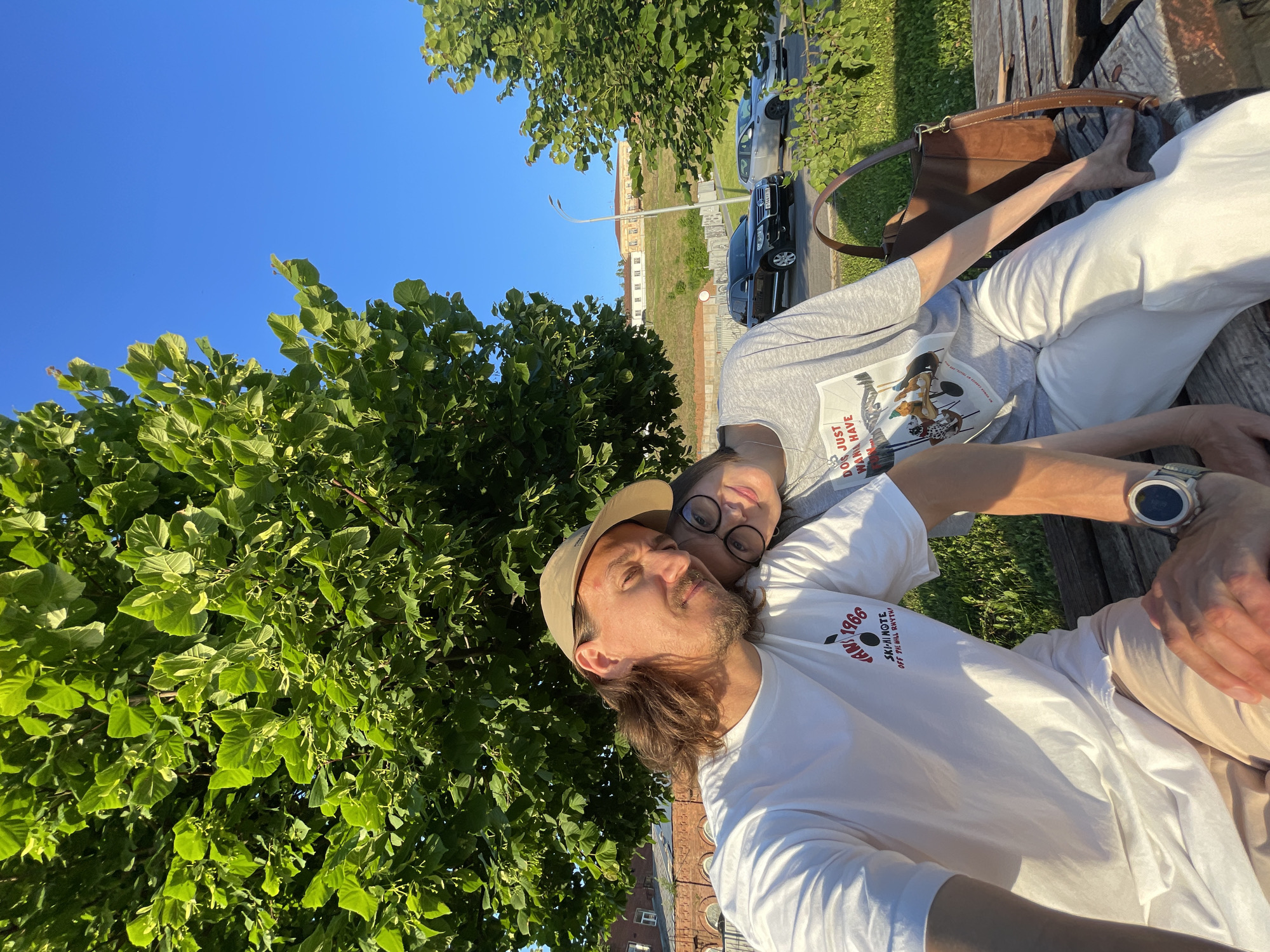 A young man and woman are sitting closely together, with the woman's head resting on the man's shoulder. They are both looking at the camera, smiling. The man is wearing a white t-shirt and a beige baseball cap, and the woman is wearing a grey t-shirt and glasses. They are sitting on a wooden bench under a large tree with green leaves. In the background, there is a street with cars and some buildings on a hill. The sky is clear and blue.