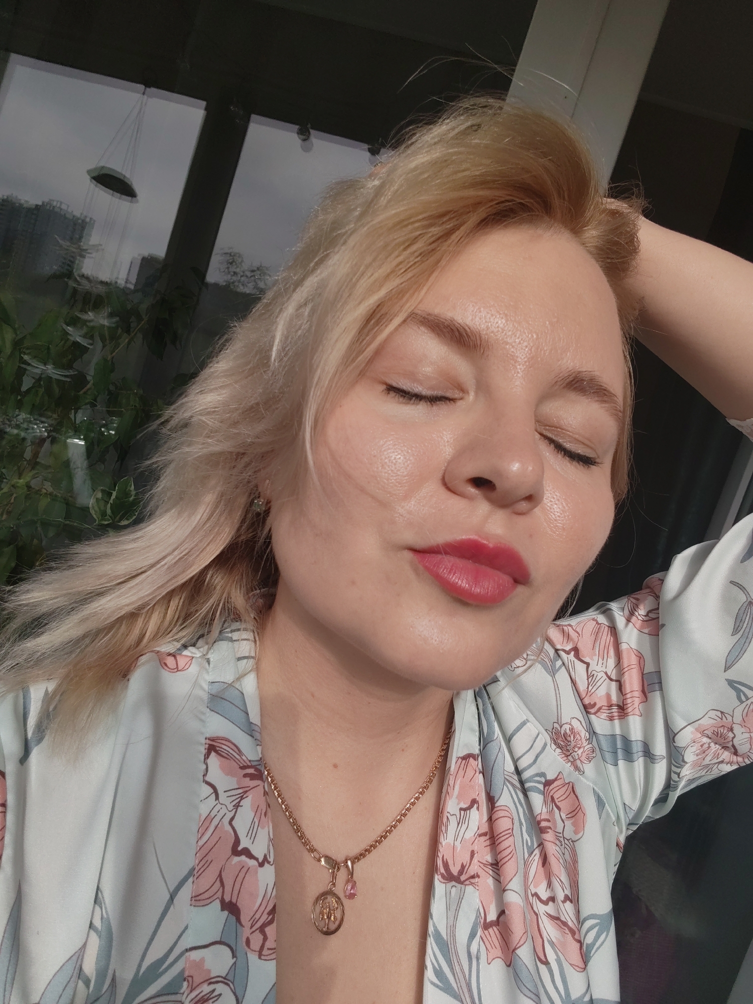 A woman with blonde hair and closed eyes is indoors, wearing a floral robe and gold necklace. Her left hand is positioned on the back of her head. Outside the window is a cityscape, suggesting an urban setting. A hanging decorative piece and plants are visible inside the building. The scene appears to be in a modern apartment or office in Izhevsk, Russia, given the architectural style of the visible buildings outside.