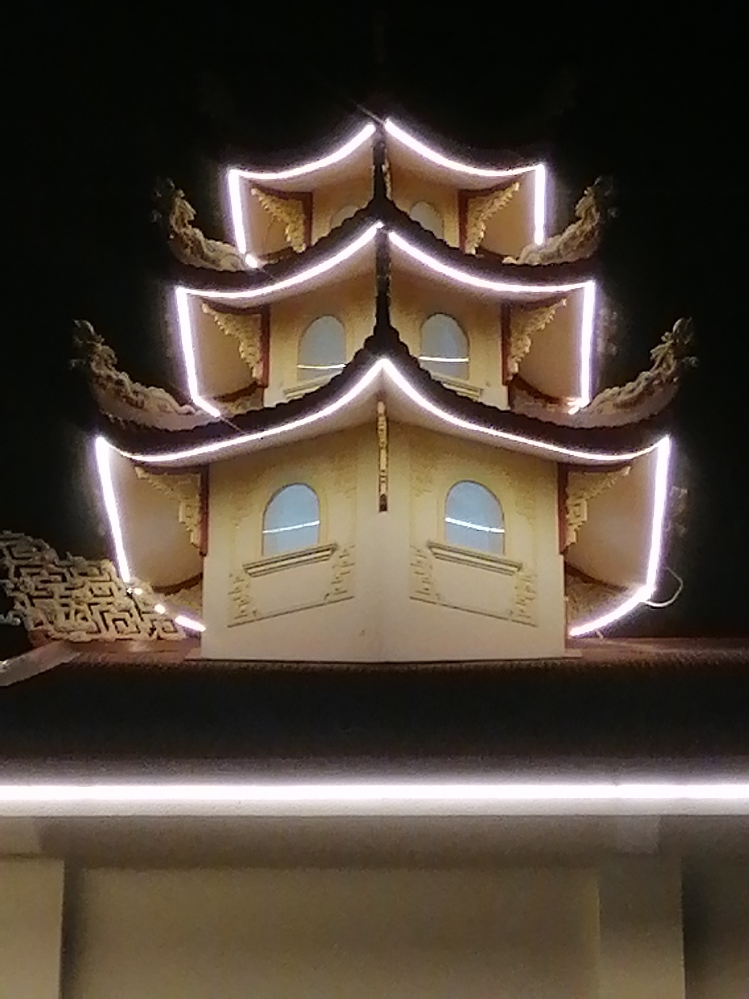 Captured at night, the image focuses on a portion of a pagoda-style structure, potentially located in the city of Da Lat, Vietnam. The structure, lit with bright white LED trim, is tiered with overlapping roofs characteristic of Asian temple architecture. Each tier is embellished with decorative elements such as curved roof edges and possibly dragon or gargoyle figures at the corners. Windows with arching tops are visible on at least two levels. The background is dark, suggesting a night scene. The image appears to be taken from a low angle, looking upward at the pagoda segment.