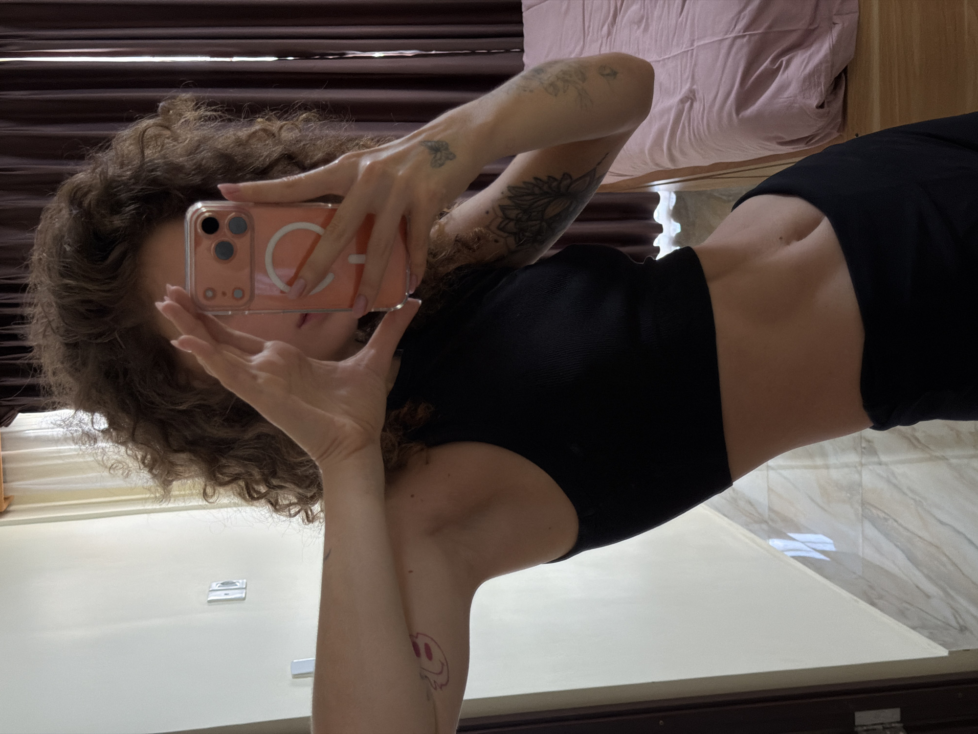 The media file is a mirror selfie taken by a person in an indoor setting. The individual, who appears to be female, is positioned centrally, holding an iPhone with a light pink case up to obscure most of their face. Their dark, voluminous curly hair frames the visible part of their head. They are wearing a black sleeveless top that exposes a slender midriff and black bottoms.
Several tattoos are visible on their arms: a large intricate mandala pattern on the right forearm, a small butterfly on the back of the right hand, and a distinct melting pink smiley face on the left upper arm. Their fingernails are painted light pink. The phone case features a prominent white letter "A" and a circular magnetic element, possibly indicating a MagSafe compatible design.
The background, reflected in the mirror, shows elements of the room. To the person's right, a bed with a light pink or beige duvet and a dark wooden headboard is visible. Behind the person's head, dark brown striped curtains or blinds can be seen. The lower portion of the image shows light-colored walls and a tiled floor with a light marble-like pattern. The lighting is ambient, characteristic of an indoor environment. The image is taken in what is noted as North Kuta, Indonesia, though no specific visual cues within the image confirm this location.