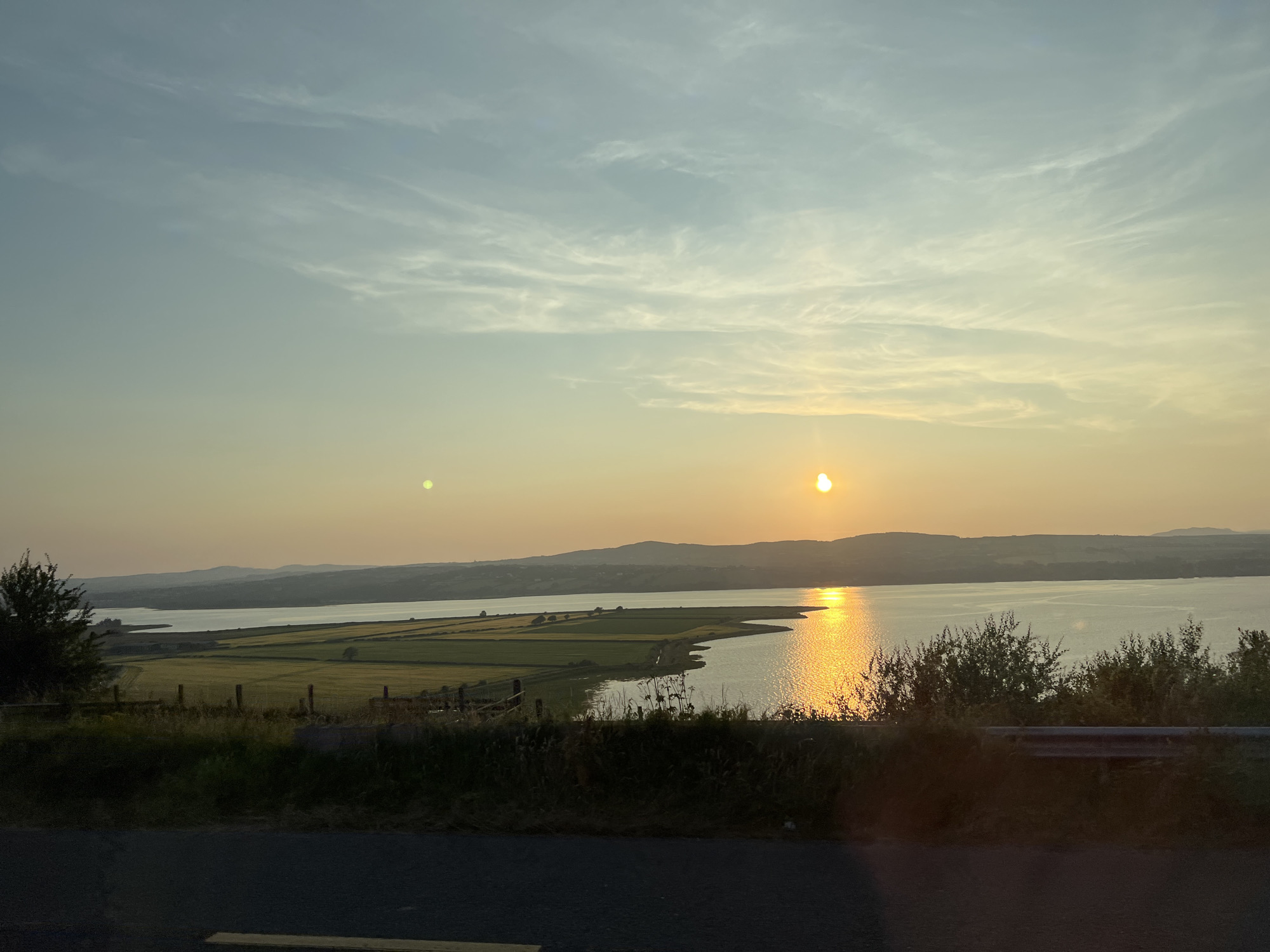 This image captures a tranquil sunset over a body of water, likely Lough Swilly, near Letterkenny, Ireland. The sky is a gradient of soft blues and yellows, with wispy clouds scattered across the expanse. The sun, a bright orb, is low on the horizon, casting a shimmering golden path across the water. In the foreground, a dark asphalt road runs horizontally across the bottom of the frame, partially obscuring a grassy embankment and a low metal barrier. Beyond the embankment, fields of green and yellow crops are visible, bordered by a wooden fence. Further in the distance, rolling hills form a soft silhouette against the horizon. The overall impression is one of peace and natural beauty, characteristic of the Irish countryside.