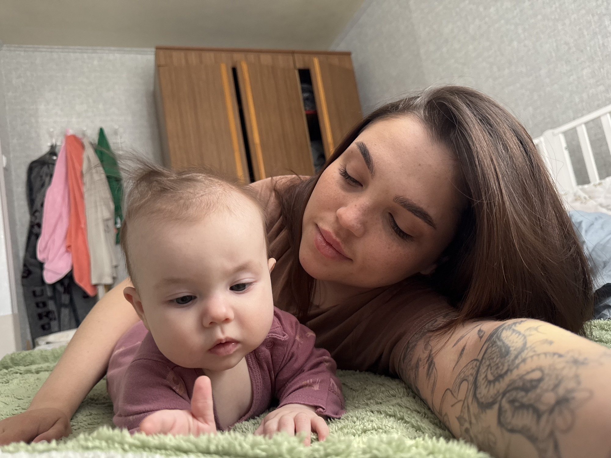 A woman and a baby are lying on a green blanket. The woman is fair-skinned with brown hair, freckles, and a tattoo on her left arm. She is wearing a brown shirt. The baby is lying on its stomach and is wearing a long-sleeved, dark pink onesie. The baby has fair skin and some hair sticking straight up. In the background is a wooden wardrobe and a clothes rack with a few garments hanging on it. The location is likely indoors, possibly a home. There is no information to confirm or deny this photo was taken in Bataysk, Russia.