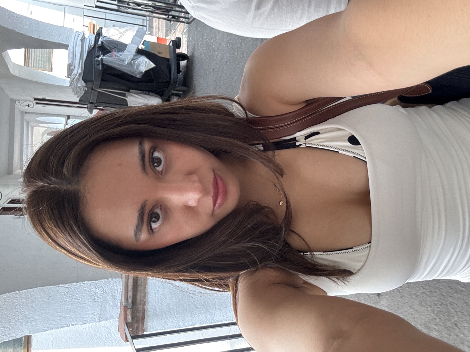 A woman with brown hair takes a selfie indoors. She wears a white tank top with a brown strap purse over her shoulder. The background shows a building exterior with arched windows and a cleaning cart in the distance. The location is inferred to be in Cocoyoc, Mexico, based on external information, although no identifying landmarks are visible in the image itself.