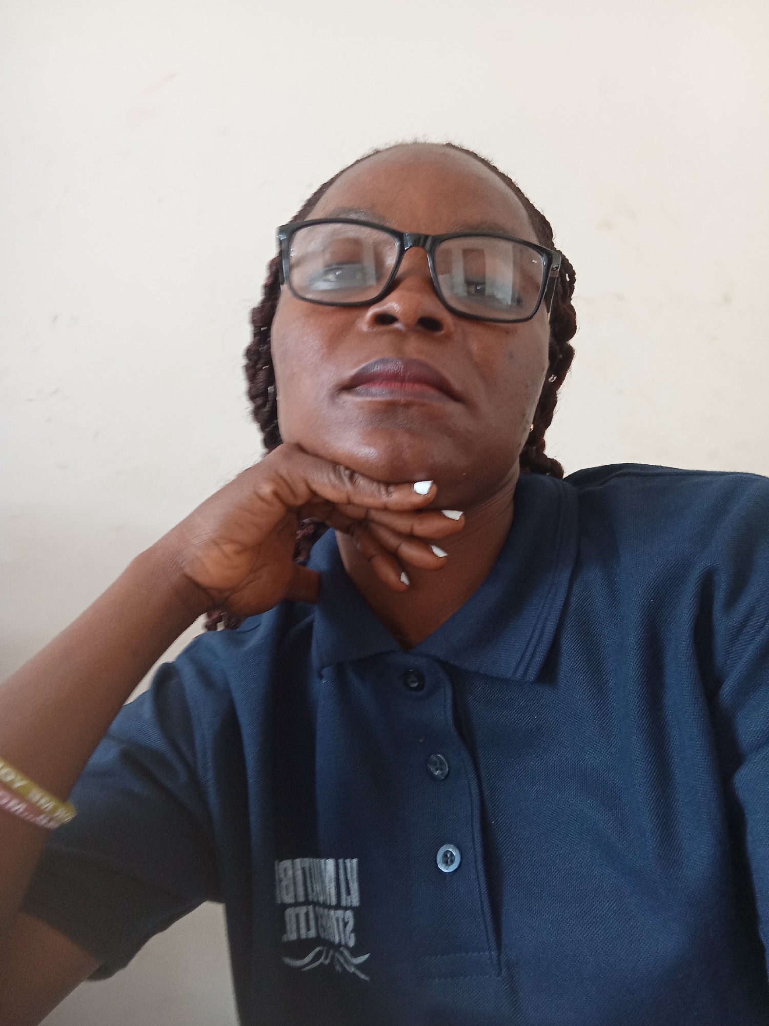 A medium shot shows a Black woman wearing glasses and a dark blue polo shirt with a logo on the left side of the chest. She is resting her chin on her right hand, with her fingers splayed and pointing upwards. Her fingernails are painted white. Her left arm is visible, resting on her chest, and she is wearing a yellow and pink bracelet on her wrist. The background is a plain, light-colored wall. The image appears to have been taken indoors, in a casual setting.