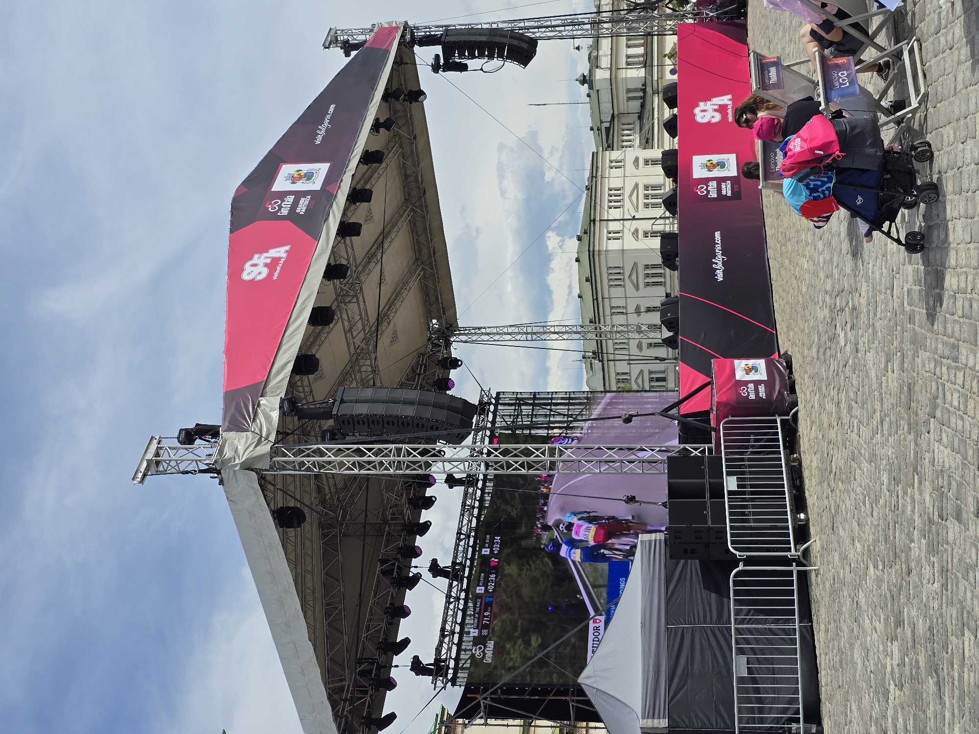The image shows a large outdoor event stage in Sofia, Bulgaria. The stage is adorned with pink and black banners displaying "visitsofia.bg" and "visitbulgaria.com" alongside the "Giro d'Italia" logo. A large LED screen displays a live broadcast of a cycling race. Various lights and speaker systems are mounted on the stage structure. People are visible on the right side of the image, including a woman with a stroller. Metal barricades are present. The backdrop features a large white building, characteristic of Sofia's architecture. The ground is paved with stone blocks.