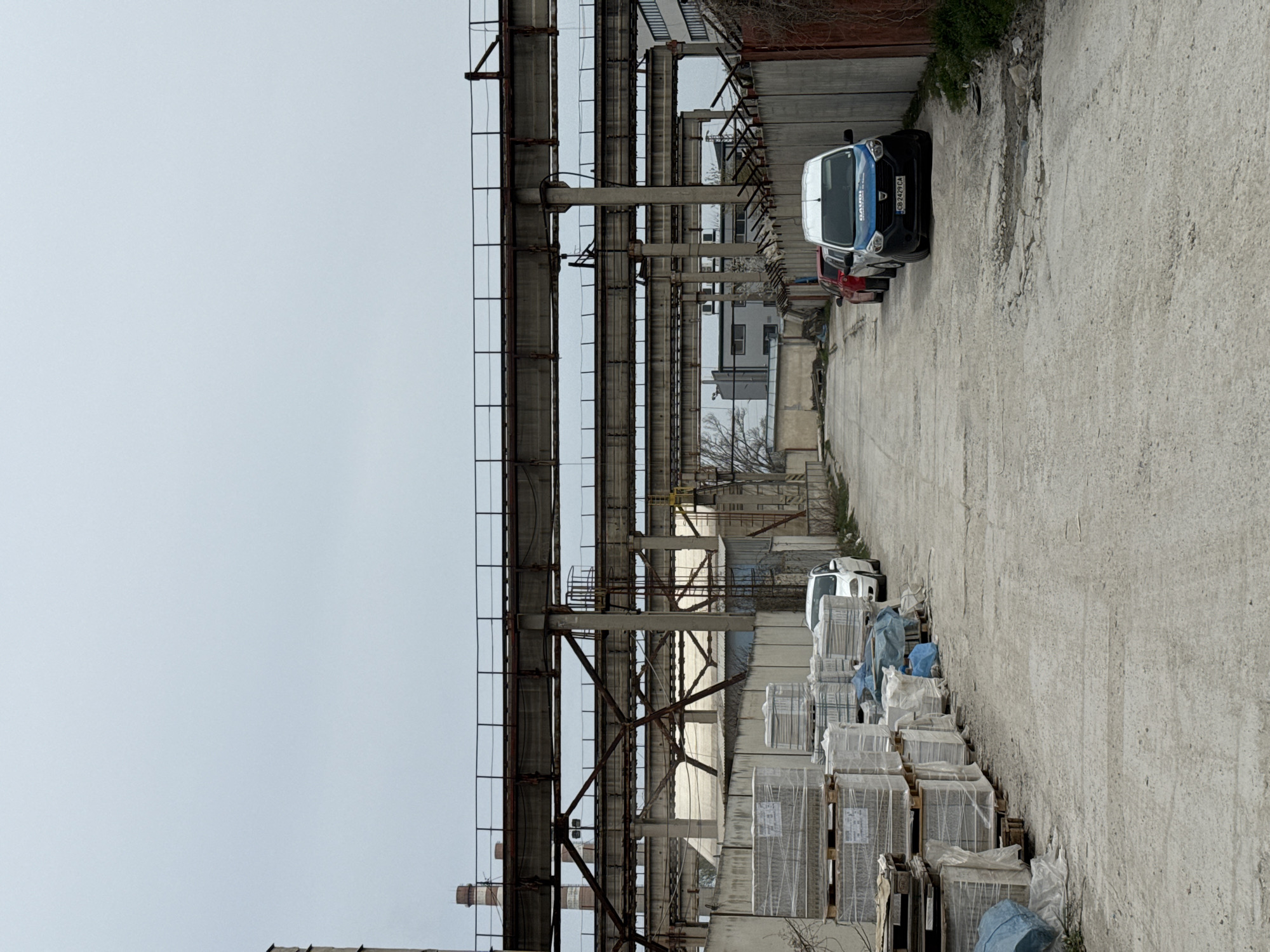 The scene is an industrial area located in Busmantsi, Bulgaria. The foreground features stacks of packaged materials on wooden pallets. To the left, a large, overhead metal structure dominates the view. In the background, a narrow road is flanked by concrete walls and industrial buildings. Three vehicles are parked along the road. The sky is overcast.