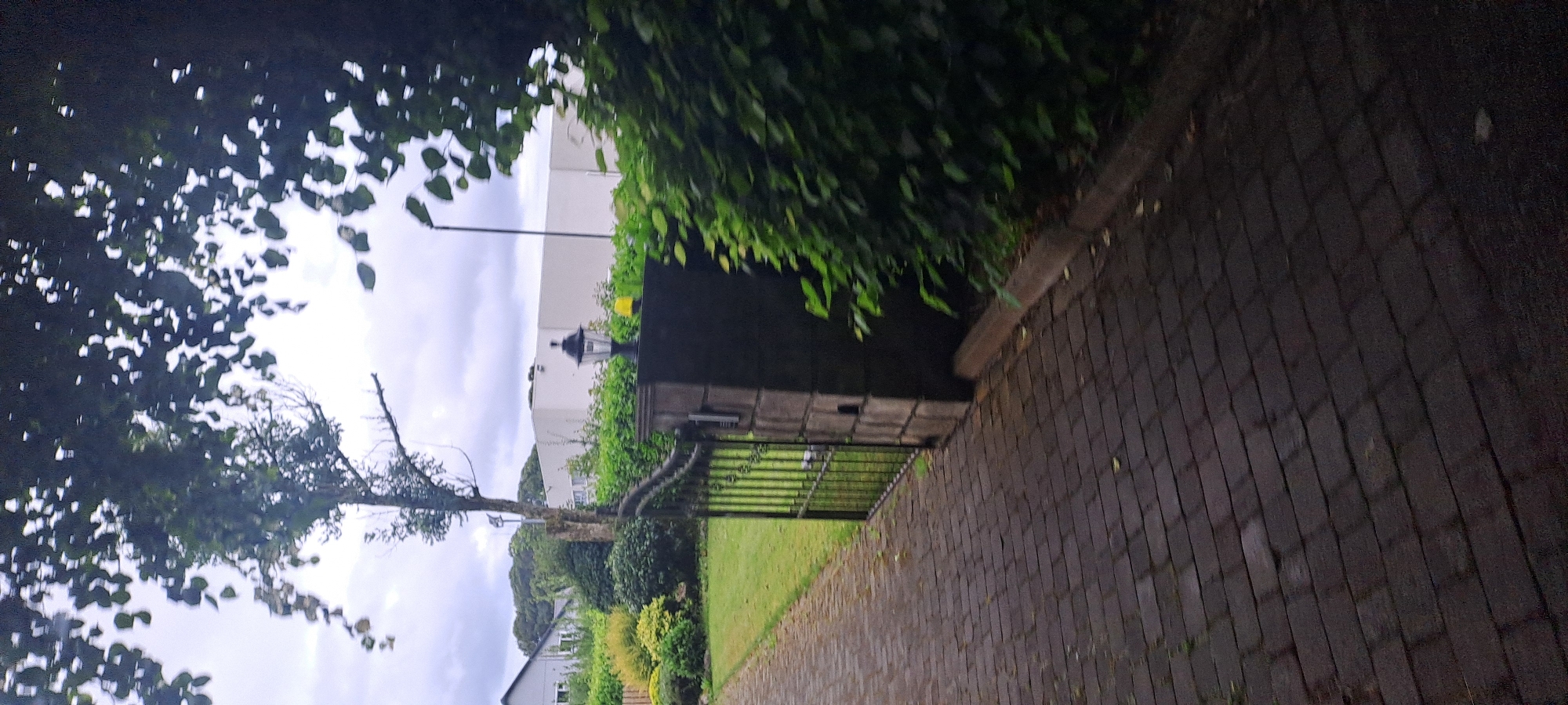 The image shows a paved driveway leading to an open black metal gate, part of a garden gate. The gate is flanked by a dark grey stone pillar on the right, topped with a black lantern with a yellow light underneath. Beyond the gate and pillar, a neatly trimmed green hedge runs along the right side of the image. To the left of the gate, a well-maintained lawn is visible, with various shrubs and trees in the background. In the far background, a white building with a flat roof and windows is partially obscured by greenery, and to the left, a residential house with a grey roof and white walls can be seen. Overcast sky with some tree branches and leaves in the foreground partially obscure the view. The scene is outdoors and appears to be a residential property.