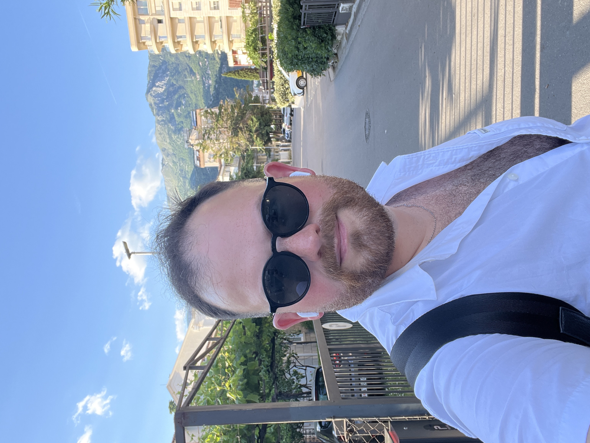 A man is taking a selfie outdoors in Budva, Montenegro. The man has light skin, dark hair, a beard, and is wearing sunglasses and a white shirt with a black strap across his chest. He also has earbuds in his ears.
Behind him is a street with cars parked along the side. Buildings are visible on the left side of the frame, along with a mountain in the background. The sky is blue with a few clouds. A metal fence with foliage is to the left of the frame. The lighting suggests it is daytime.