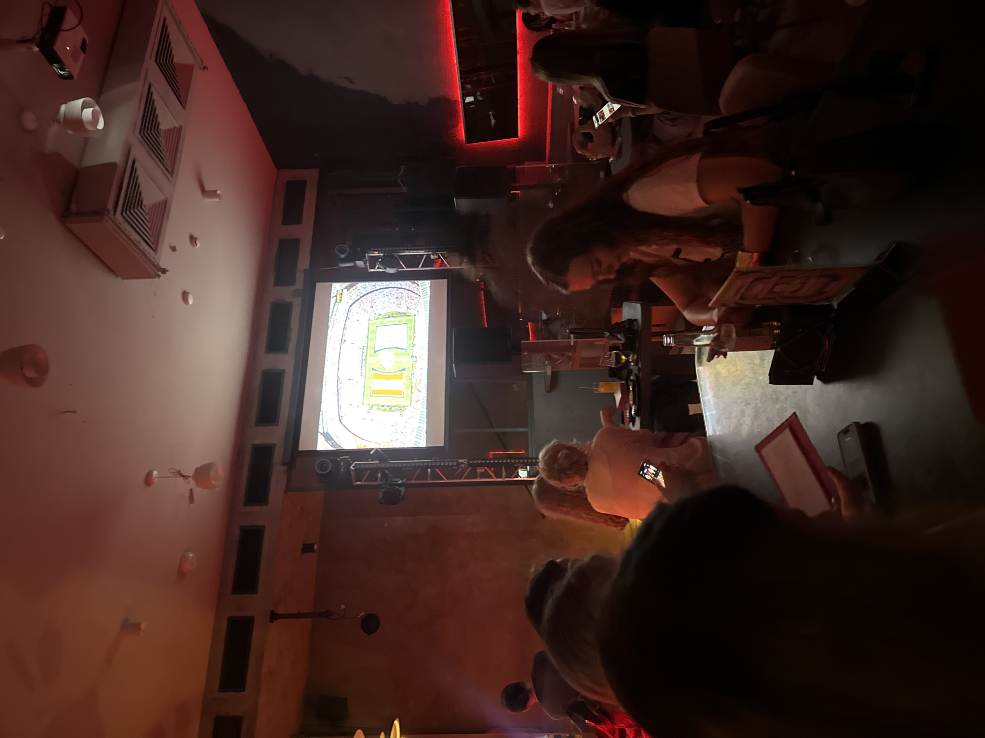 The image depicts the interior of a dimly lit establishment, possibly a bar or restaurant, where patrons are gathered to watch a sports event projected on a large screen. The screen displays a football stadium with a game in progress. The lighting is predominantly red and orange, creating a warm and somewhat moody ambiance. Several individuals are visible, some seated at tables, others standing or walking in the background. Many are looking towards the screen, engrossed in the game. One person in the foreground is holding up a smartphone, likely recording or taking a picture of the screen. Another person is looking at their phone on the table. The environment features typical bar or restaurant elements such as tables, chairs, and speakers. The overall impression is one of a social gathering focused on a shared viewing experience. The image does not provide any specific visual cues that would pinpoint its location to Moscow, Russia.