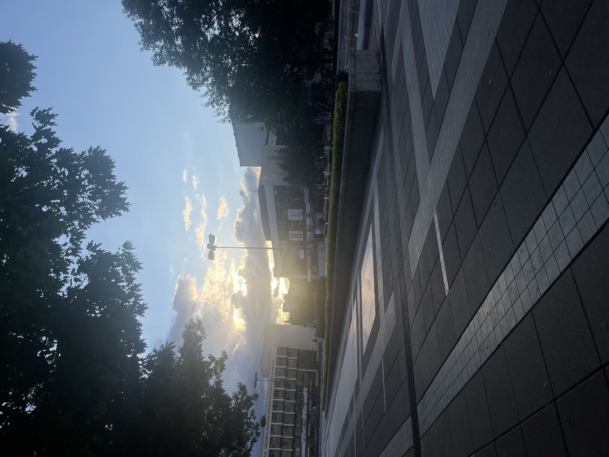 The image captures an urban outdoor scene at sunset or late afternoon in Blagoevgrad, Bulgaria. The sun, low on the horizon, casts a warm glow through scattered clouds, creating bright highlights and lens flare, particularly noticeable in the sky and reflected on the ground. The sky is predominantly blue with wispy white and gold-tinted clouds.
In the foreground and extending to the right, a wide paved area features a geometric pattern of dark grey and lighter grey/white tiles, some of which reflect the sky and sunlight. To the left, dense green trees, silhouetted against the bright sky, frame the scene.
In the midground, several modern buildings are visible. One prominent multi-story building, likely part of a university campus, bears Cyrillic text on its facade: "НЕОФИТ РИЛСКИ" (Neofit Rilski) and "ИСТОРИЧЕСКИ ФАКУЛТЕТ" (Historical Faculty), indicating it's possibly the Faculty of History at the South-West University "Neofit Rilski". Below this building, small water fountains and bushes are visible. A distinctive streetlight with multiple lamp heads stands near the buildings.
Faint figures of people can be seen near the buildings and fountains, suggesting a public space where individuals might be walking or gathered. The overall atmosphere appears peaceful and serene, enhanced by the golden hour lighting.