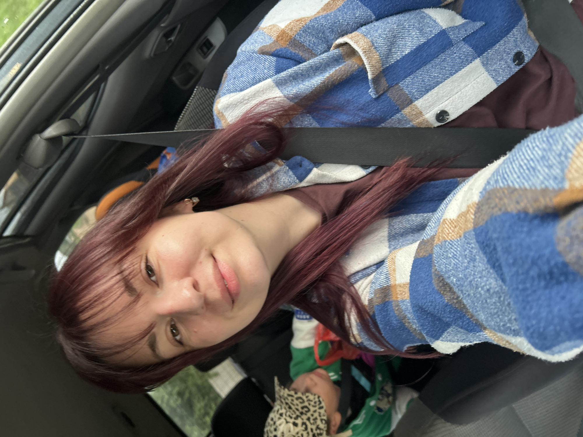 A young woman with long, dark red hair and bangs is taking a selfie in a car. She is wearing a blue, white, and tan plaid shirt over a dark reddish-brown shirt. A black seatbelt crosses her chest. Her expression is relaxed and slightly smiling. In the background, another person, possibly a child, can be seen wearing a green shirt and a leopard print hat, lying down. The setting appears to be a car interior, with a view of trees and greenery through the window, suggesting it is daytime and the weather is likely mild. The image is taken from a slightly tilted angle. There is no visible text in the image.